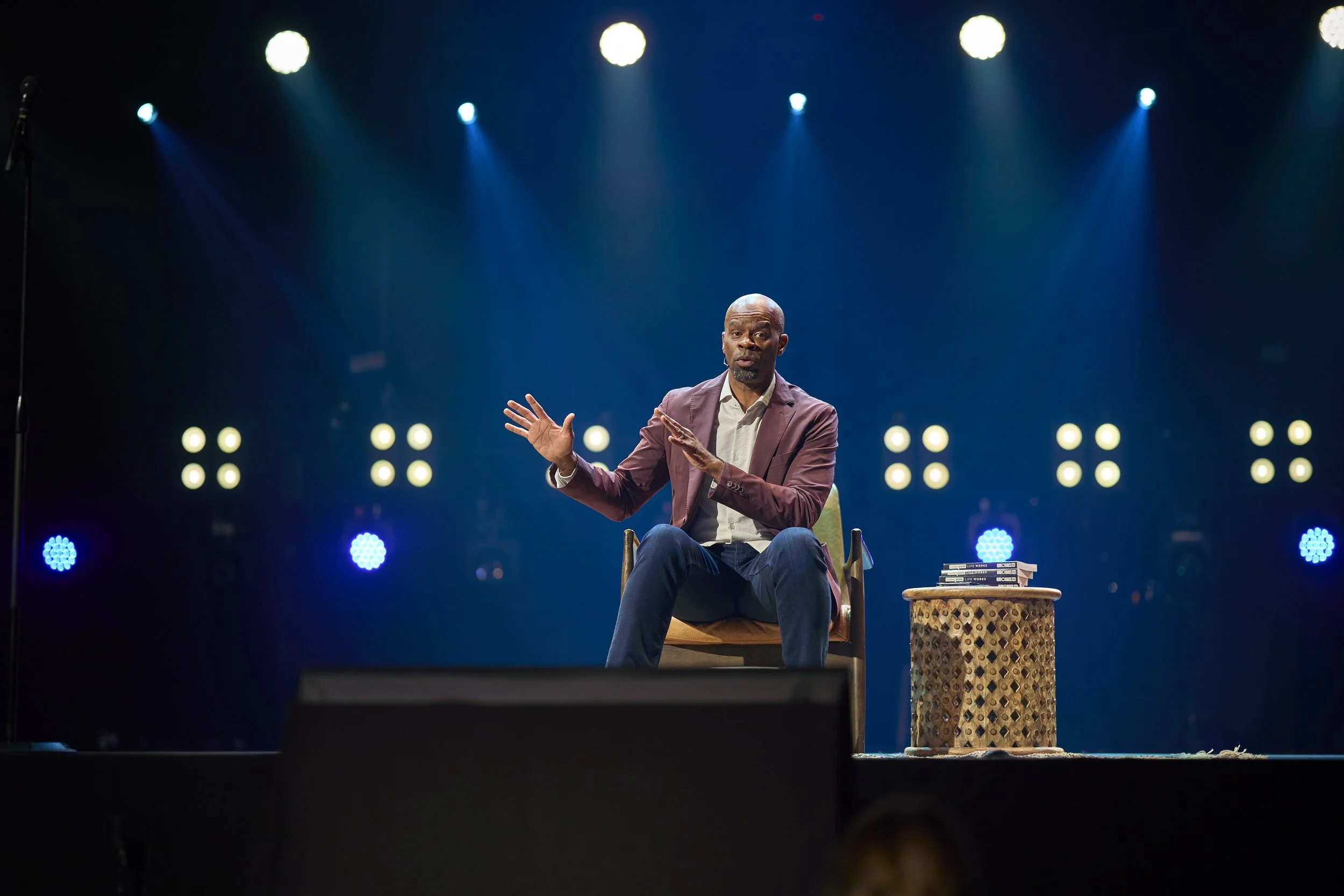 A man in a brown blazer and white shirt sitting on a stage, gesturing with his hands during a presentation. There is a small table with books and a decorative container next to him. Bright stage lights are shining from above in a dark background.