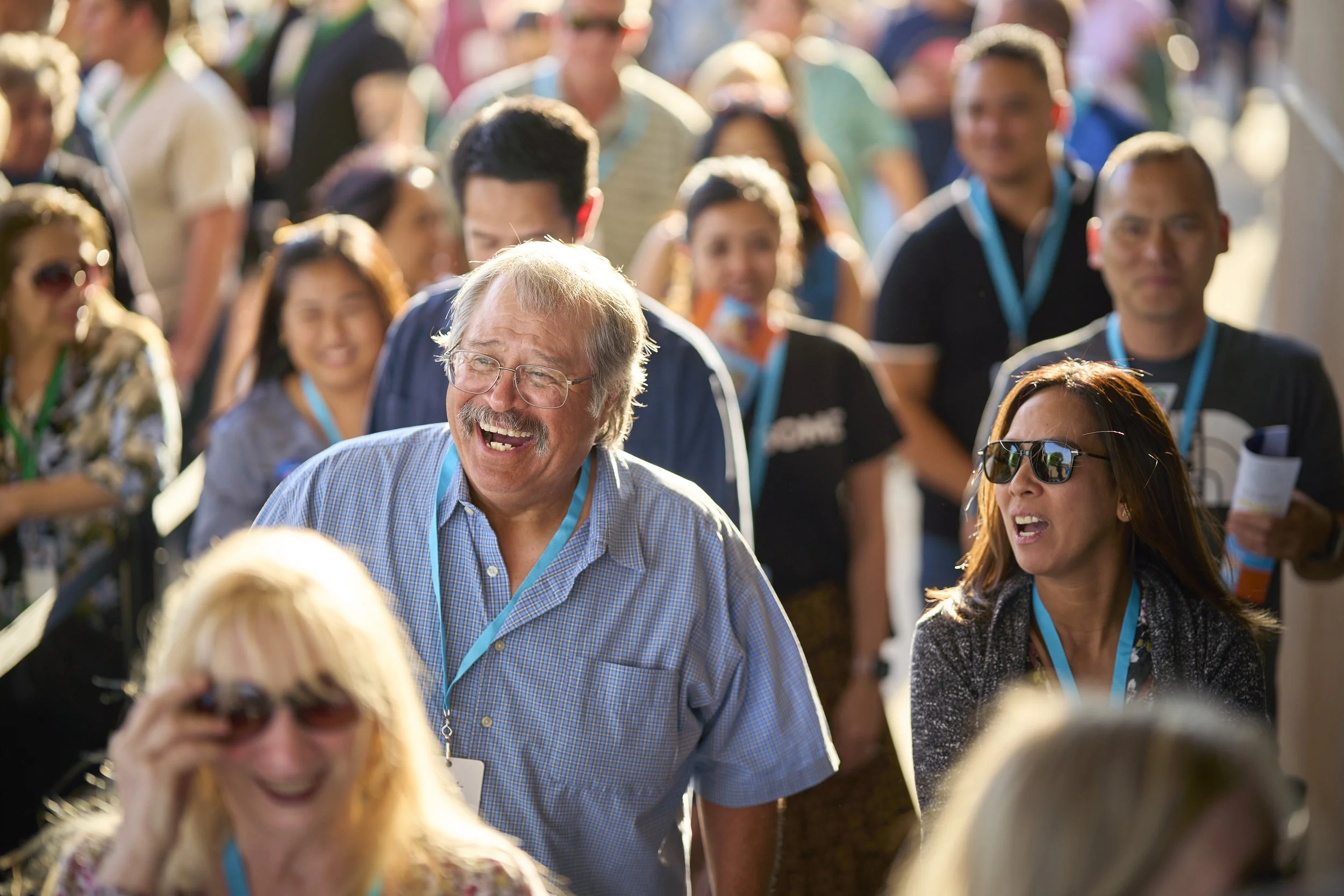 A group of people at an outdoor event. The focus is on an older man with glasses and a beard, smiling and laughing. The crowd around him appears to be engaged and having fun.
