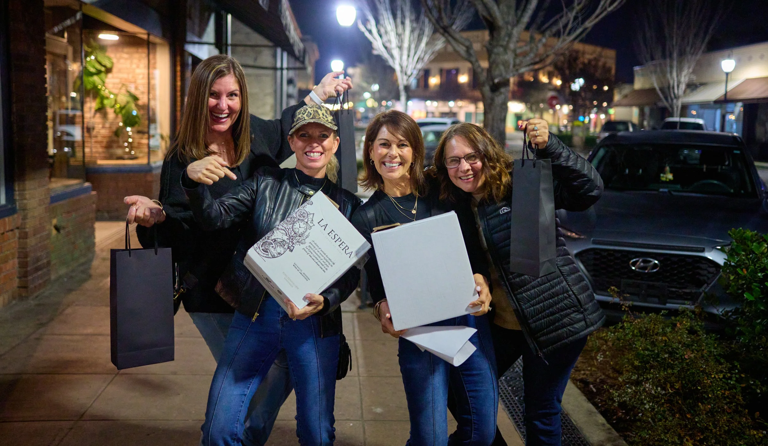 Four women standing outside at night, smiling, holding shopping bags and boxes. They are dressed casually, with two wearing glasses and one wearing a leopard print hat. Behind them, there are parked cars, trees, and illuminated storefronts.