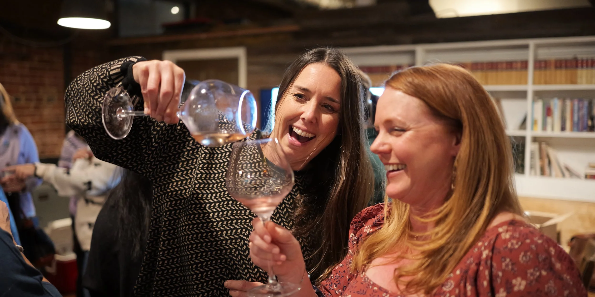 Two women smiling and clinking wine glasses at a social gathering in a cozy indoor setting with bookshelves, exposed brick, and warm lighting.