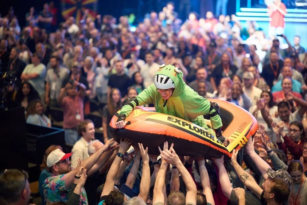 Person in a dinosaur costume riding an inflatable boat, being lifted by a crowd at a lively indoor event.