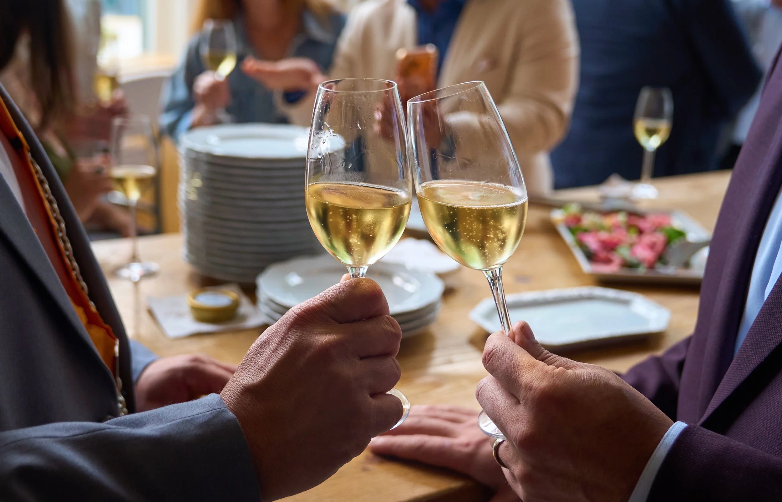 Two people clinking glasses of white wine at a social gathering, with a table of plates and food in the background.
