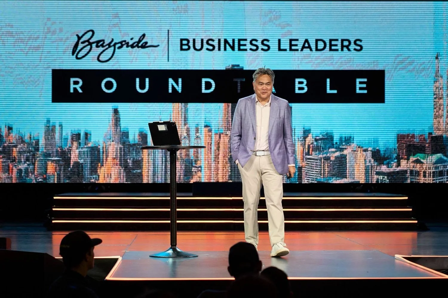 A man in a light purple blazer and cream pants standing on a stage in front of a large screen that reads 'Bayside Business Leaders Roundtable.' The stage has steps with orange lighting, and a table with a laptop and water bottles. The audience is vis