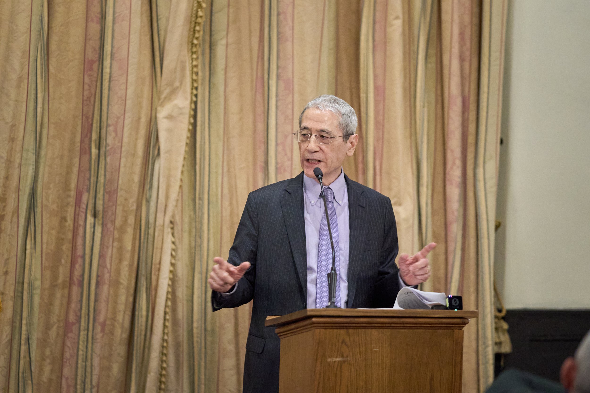 A man with glasses in a suit giving a speech from behind a wooden podium, with curtains in the background.