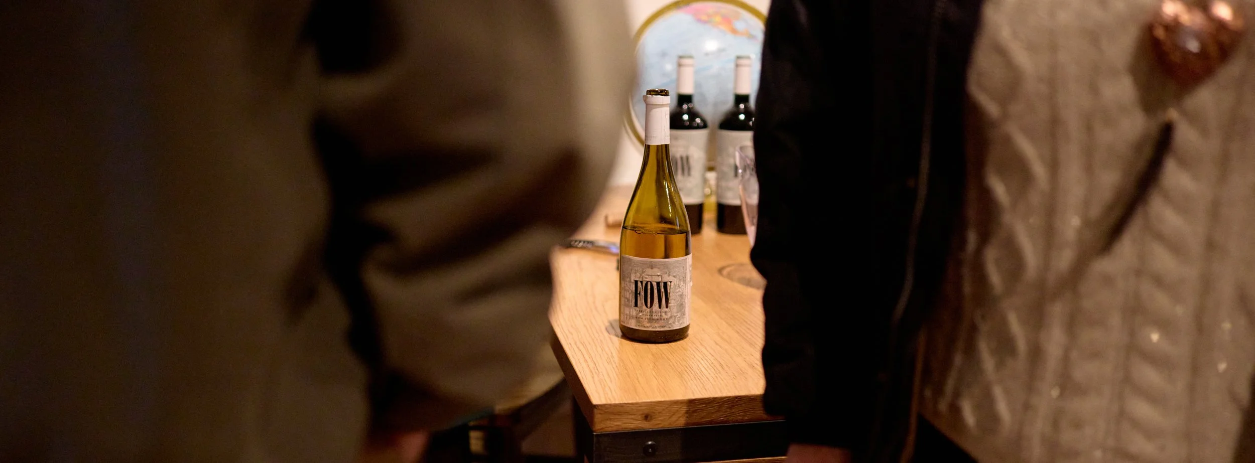 A wine bottle with a label reading 'FOW' on a wooden bar, surrounded by people at a social gathering.