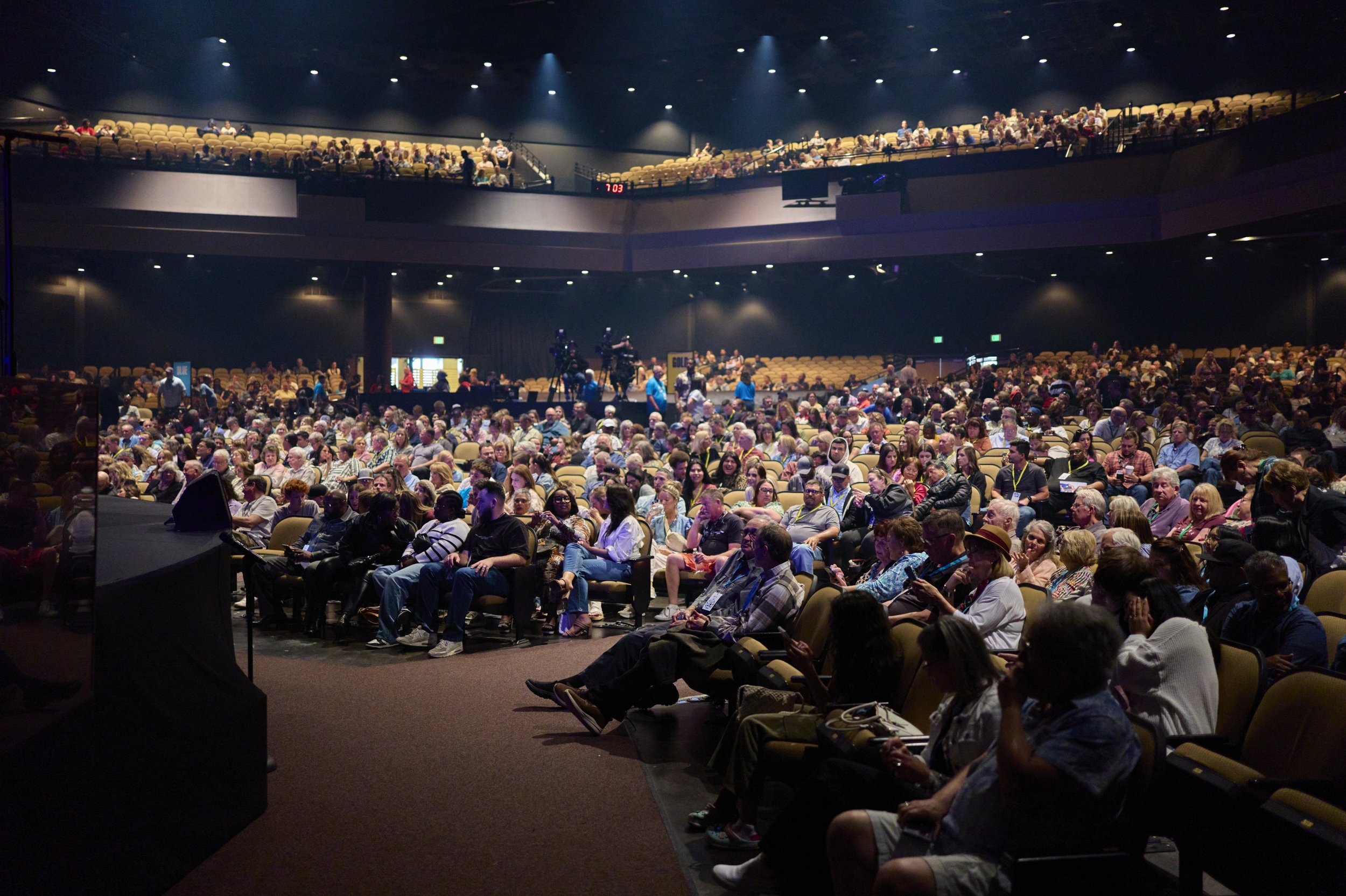 A large auditorium filled with people seated in chairs and watching a presentation or event. The stage is partially visible in the foreground with some equipment and photographers.