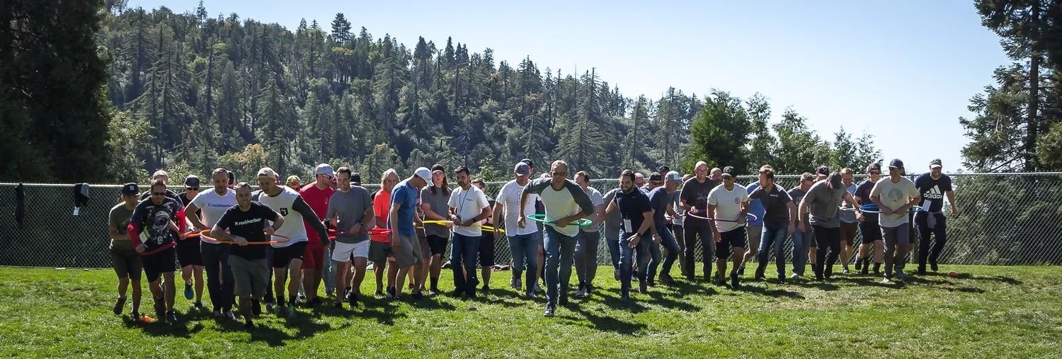 Group of people participating in a team-building activity outdoors on a grassy field, with a forested hillside in the background.