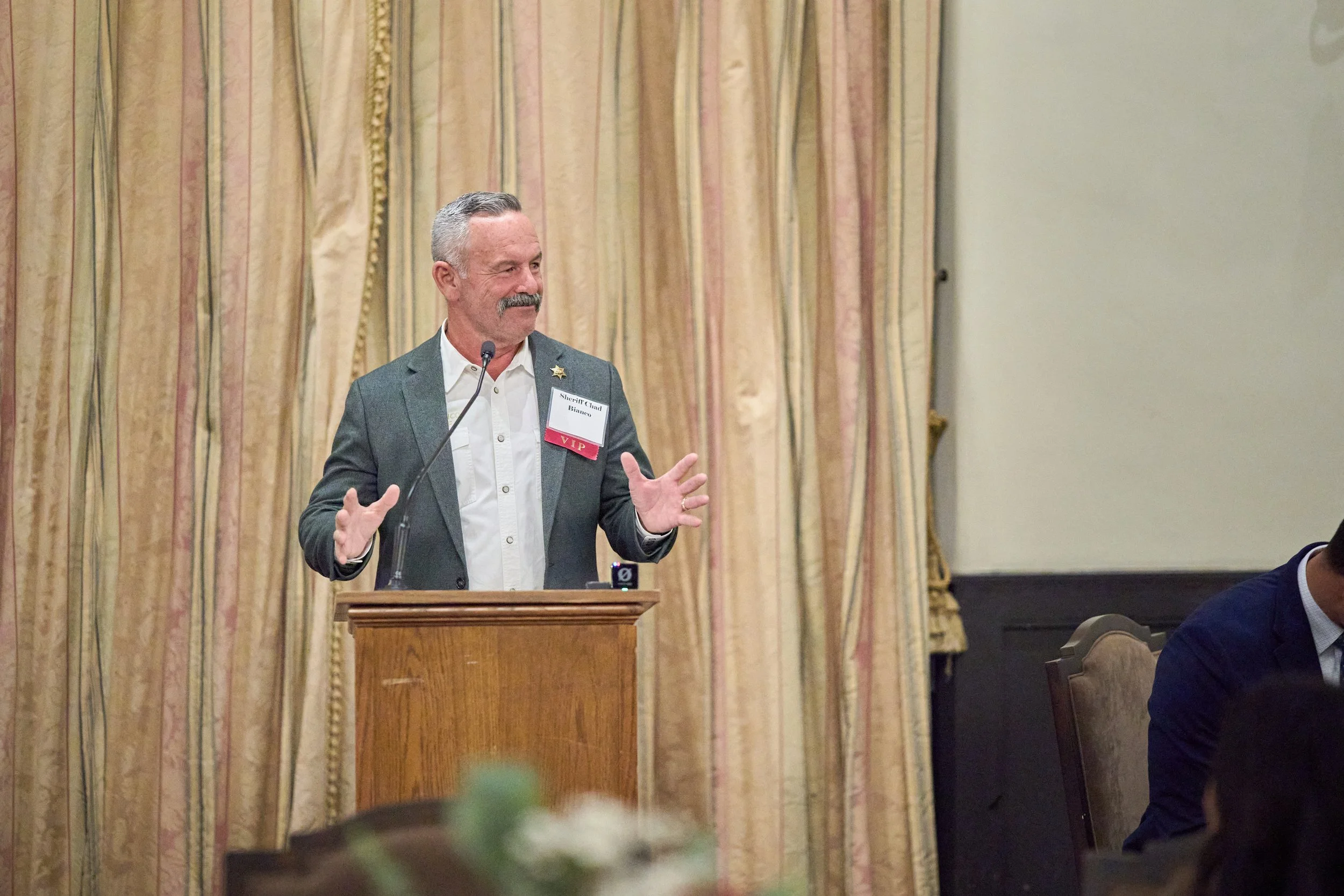 A man with a mustache and gray hair, wearing a suit jacket and a name tag, is speaking at a podium with a microphone. He is gesturing with his hands in front of a curtain with vertical stripes. There are other people seated nearby, partially visible 
