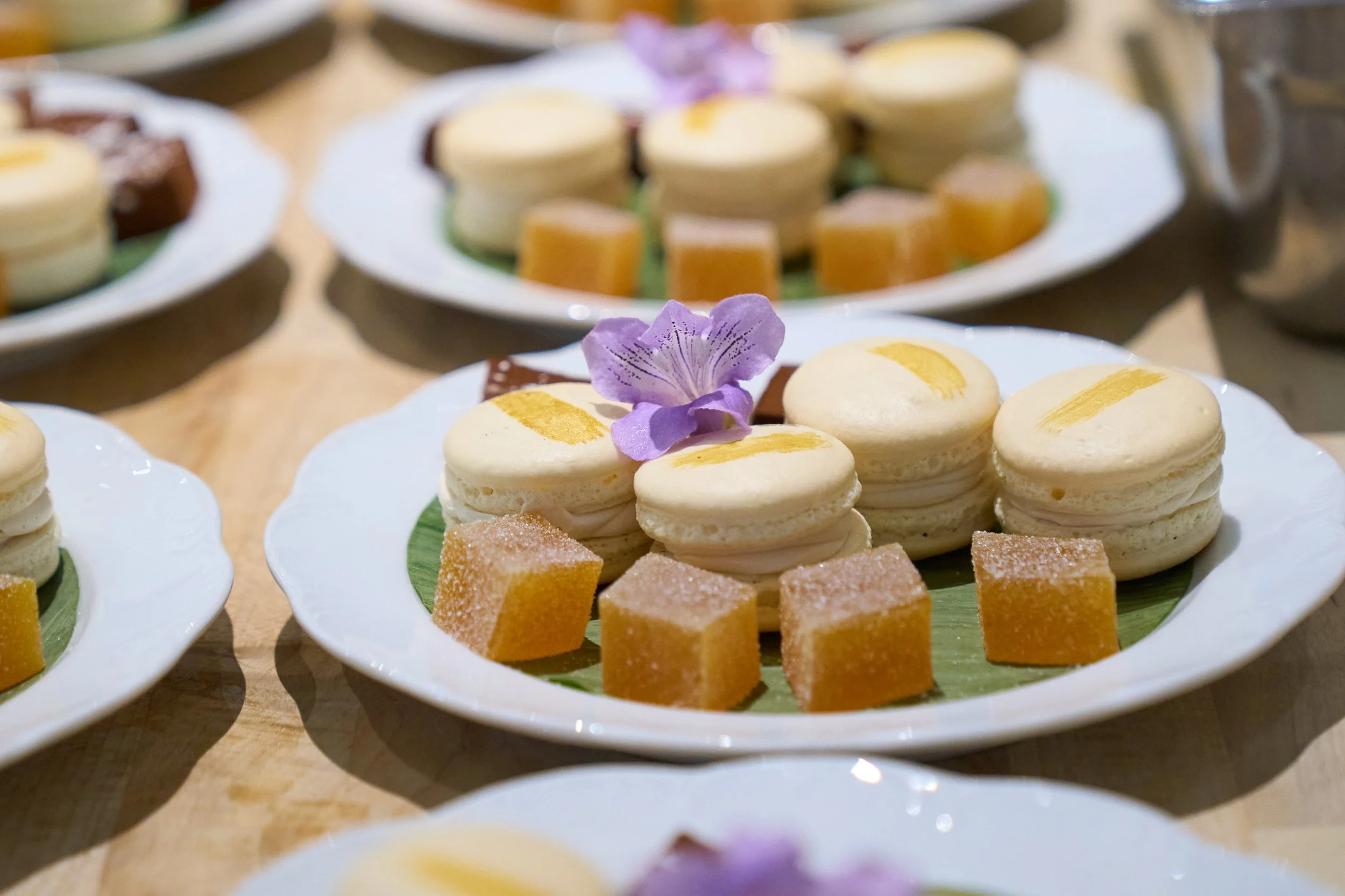 Assorted desserts on white plates, including macarons, fruit jellies, and squares, garnished with purple edible flowers.