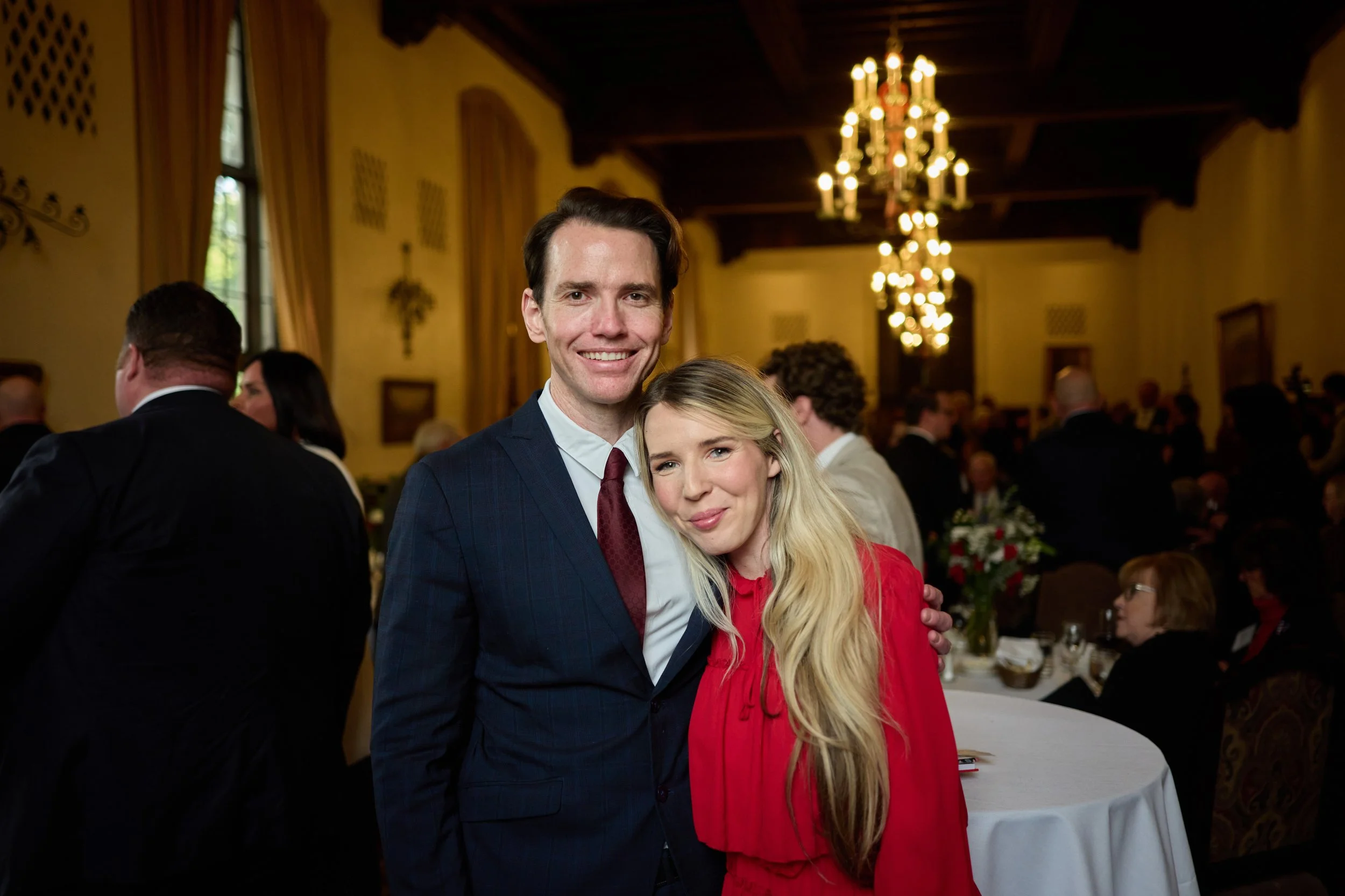 A smiling man and woman at a formal event, with the man wearing a suit and tie and the woman in a red dress, standing together in a decorated banquet hall with chandeliers and other guests in the background.