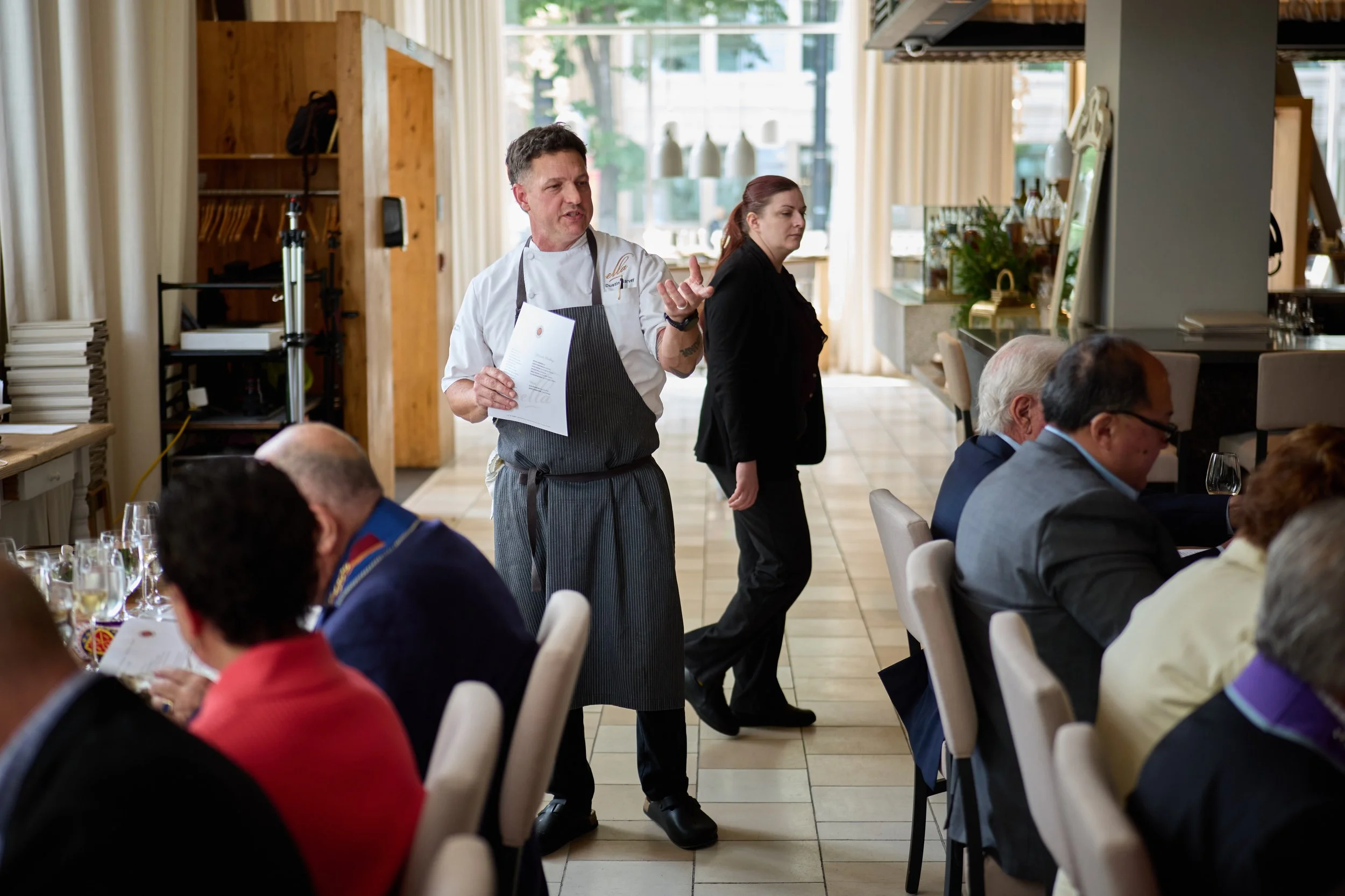 Chef in a striped apron and white chef coat holding a menu, speaking while standing in a restaurant with guests seated at tables, and a woman in black standing nearby.