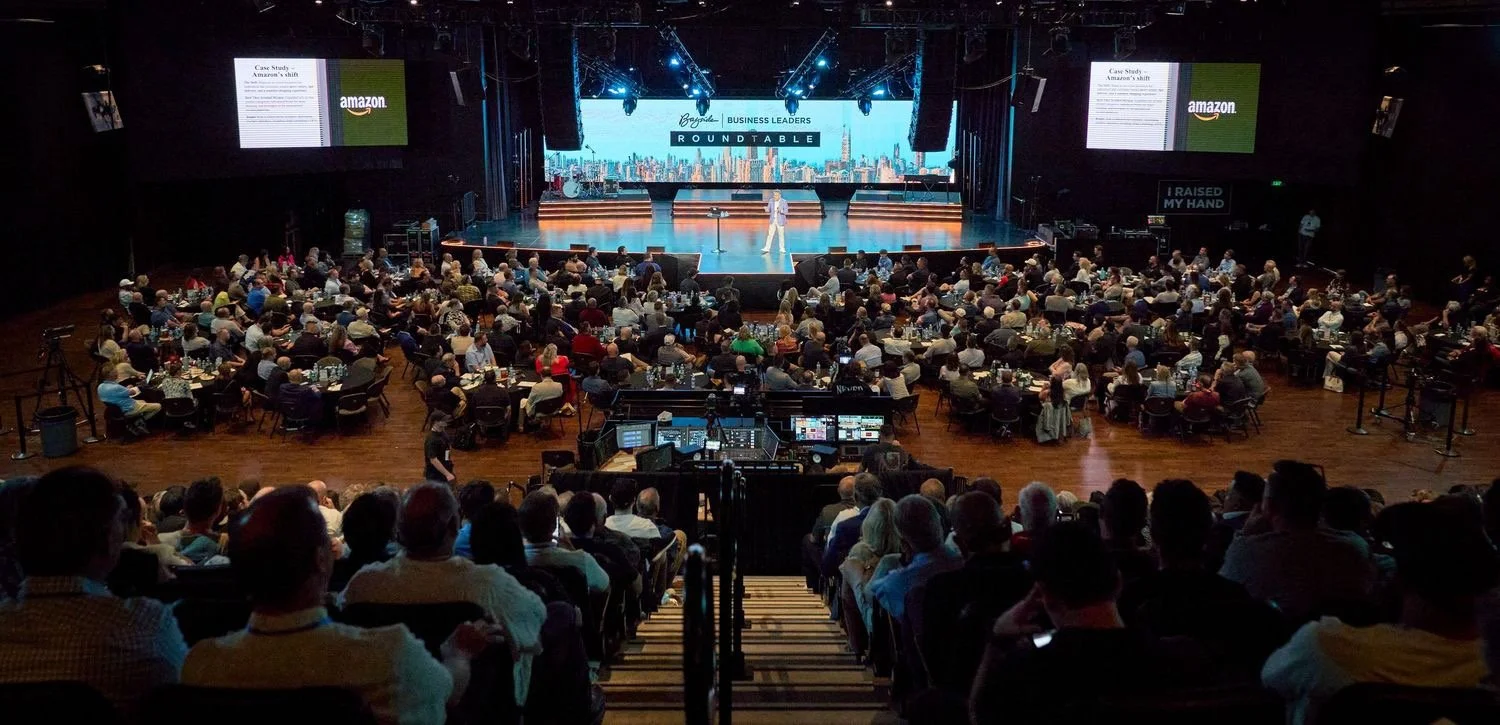 A large conference room filled with an audience watching a speaker on stage during a business event, with large screens displaying the Amazon logo and a presentation behind the speaker.