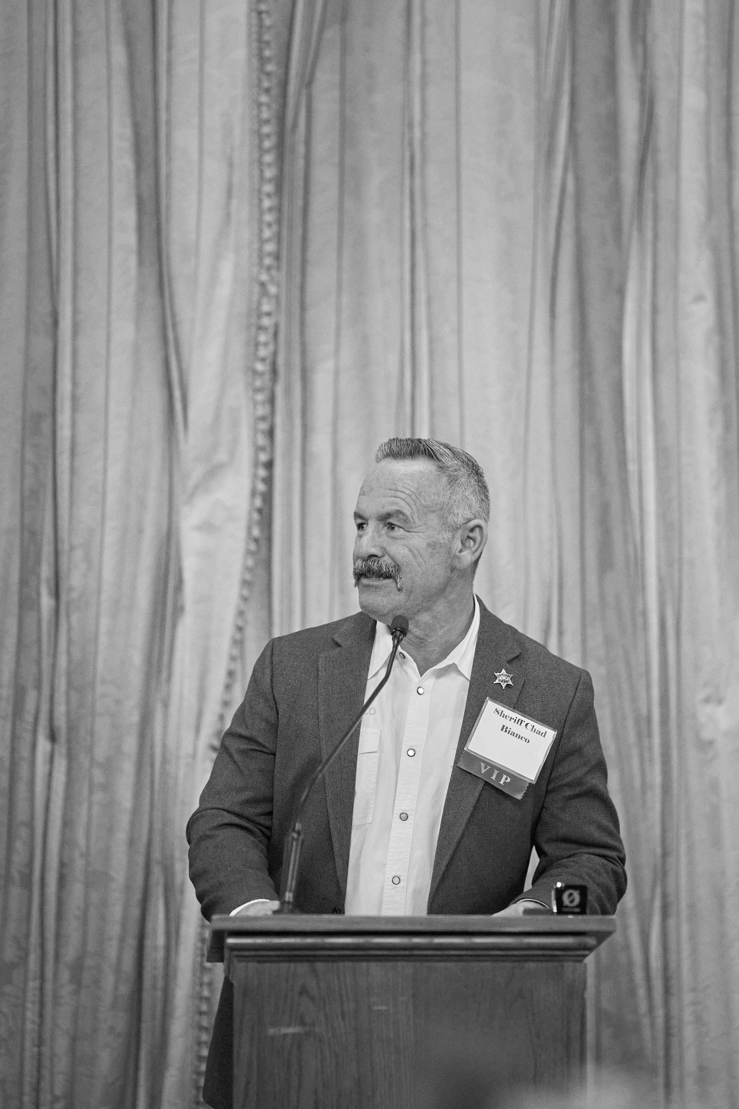 A man with a mustache and short hair, wearing a suit and white shirt, standing at a podium with a microphone, in front of patterned curtains, wearing a name tag that reads 'Sheriff Chad Bianco' and 'VIP'.