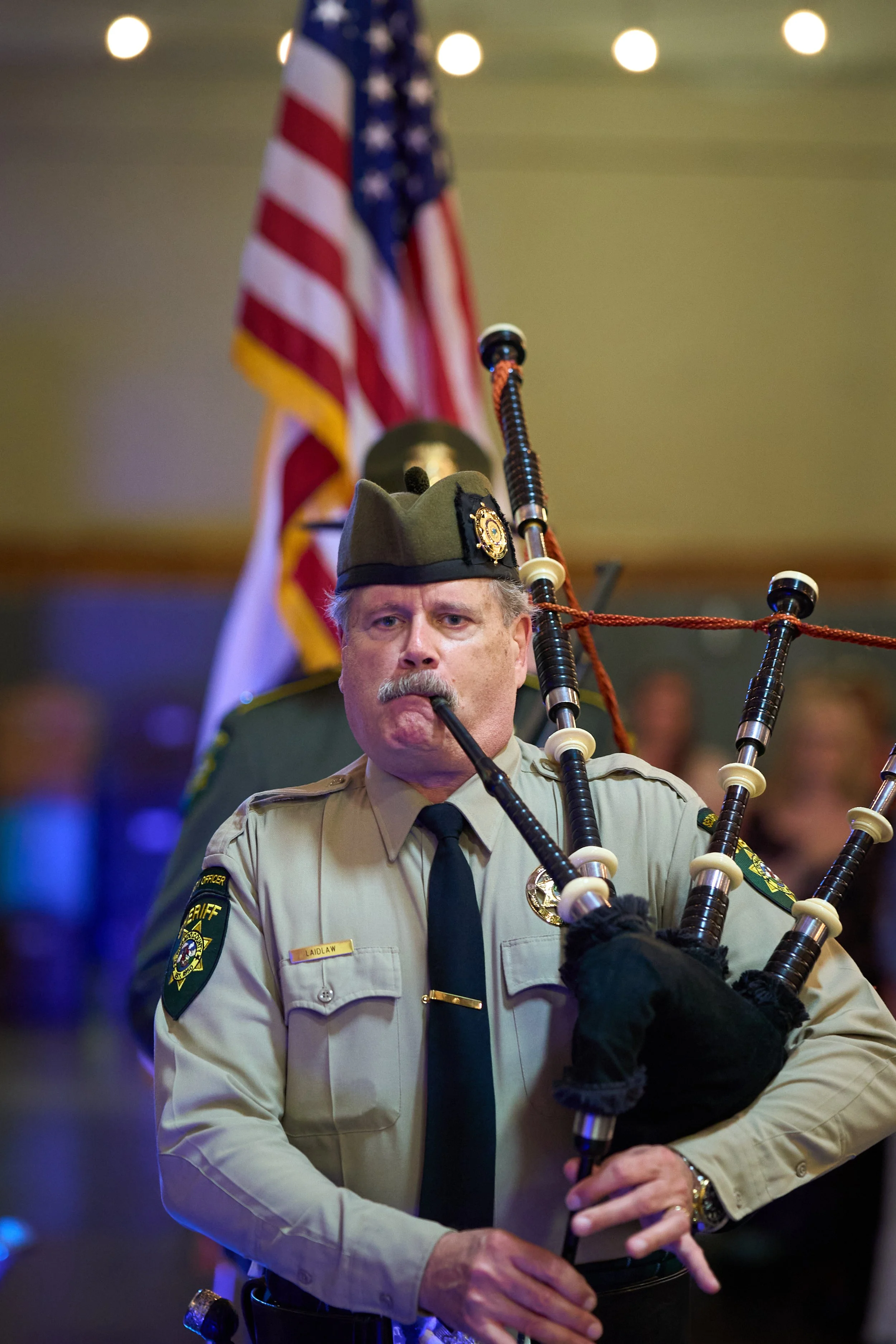 A sheriff in uniform playing bagpipes indoors with an American flag in the background.