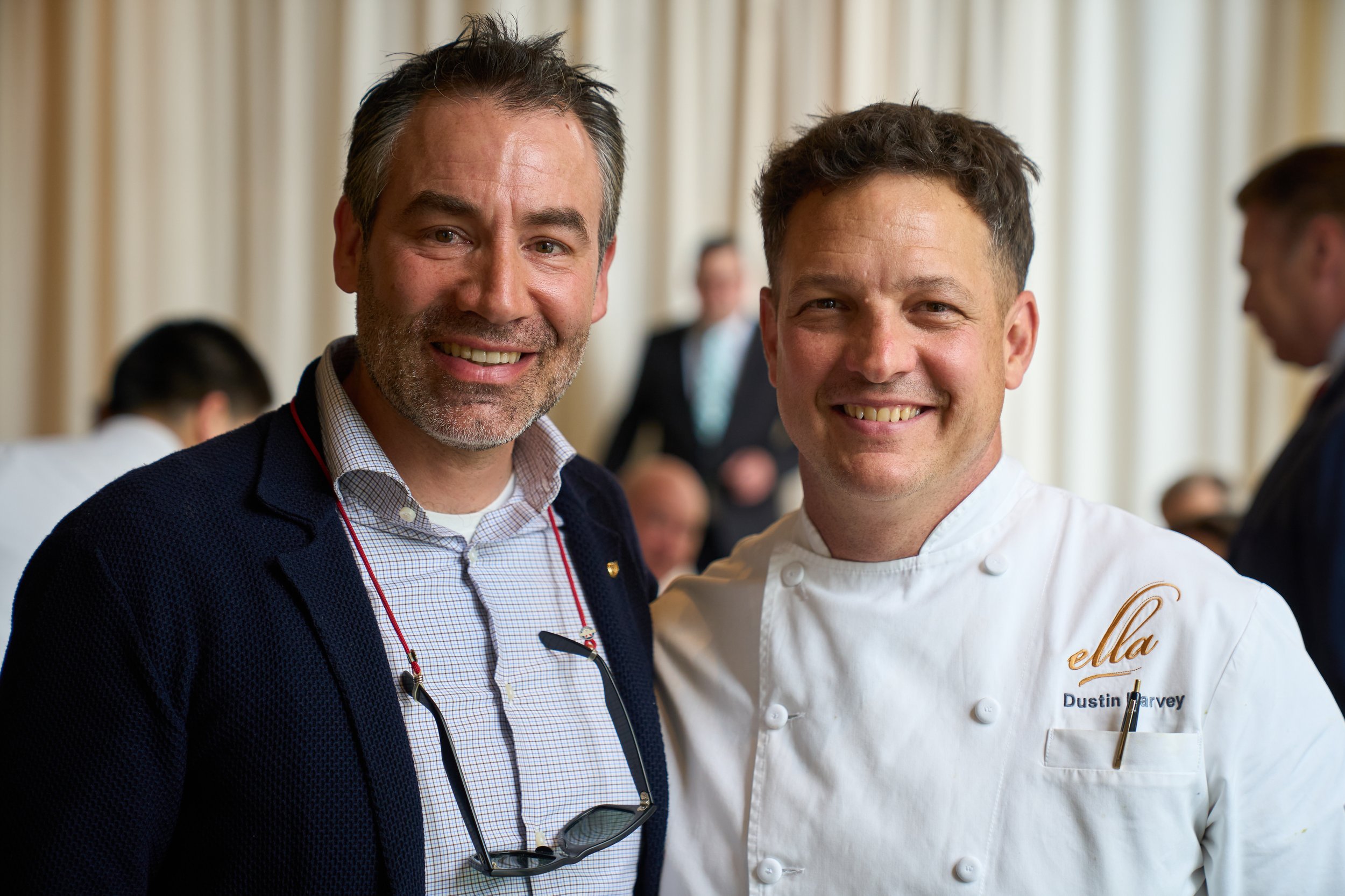 Two smiling men, one in a dark jacket with glasses hanging from his shirt, and the other in a white chef's coat with "ella" embroidered on it, standing close together at an event with a blurred background of other people.