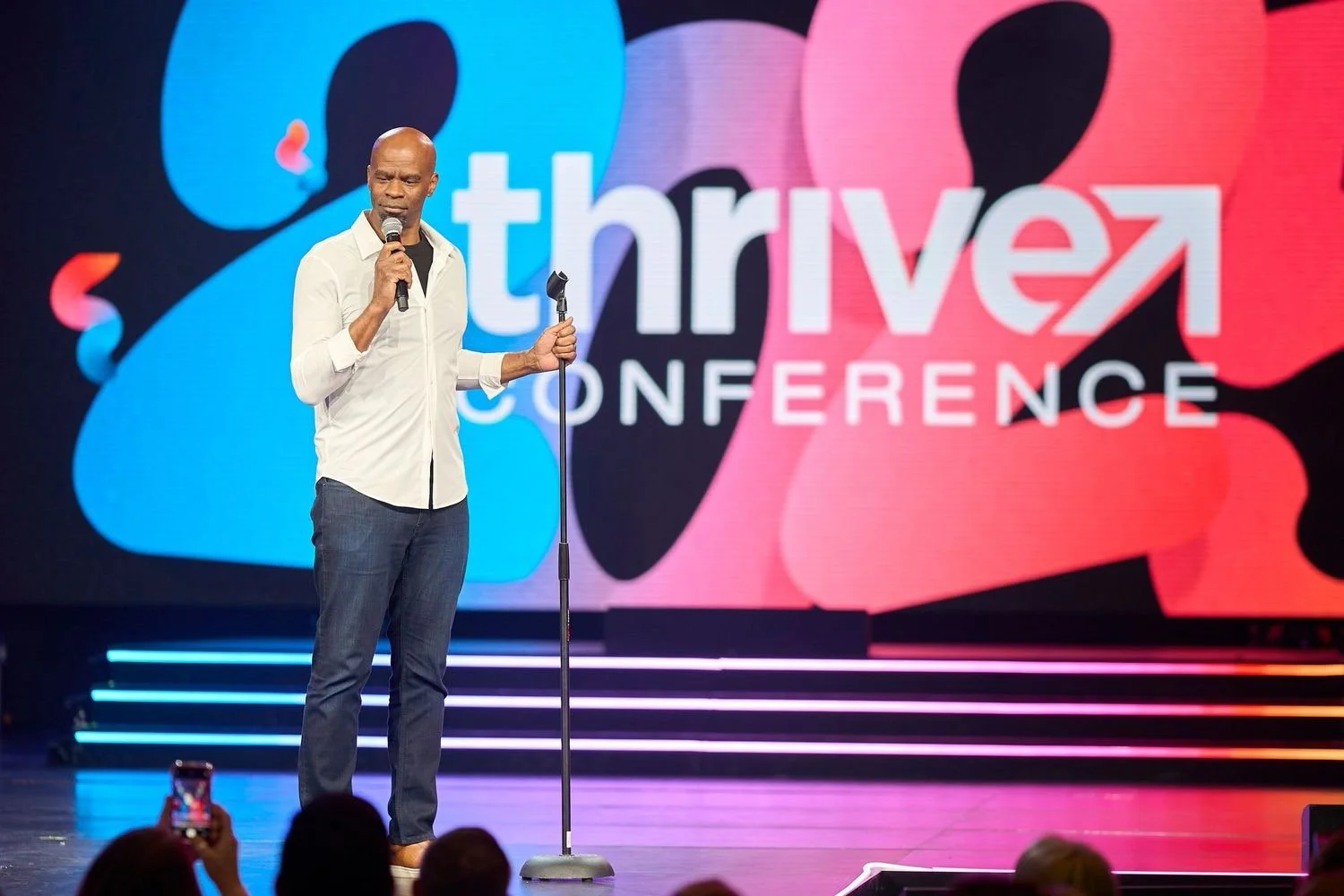 A man standing on stage at the Thrive Conference holding a microphone while speaking, with a colorful large screen behind him displaying the words "Thrive Conference" in white text, with large blue and pink abstract shapes.