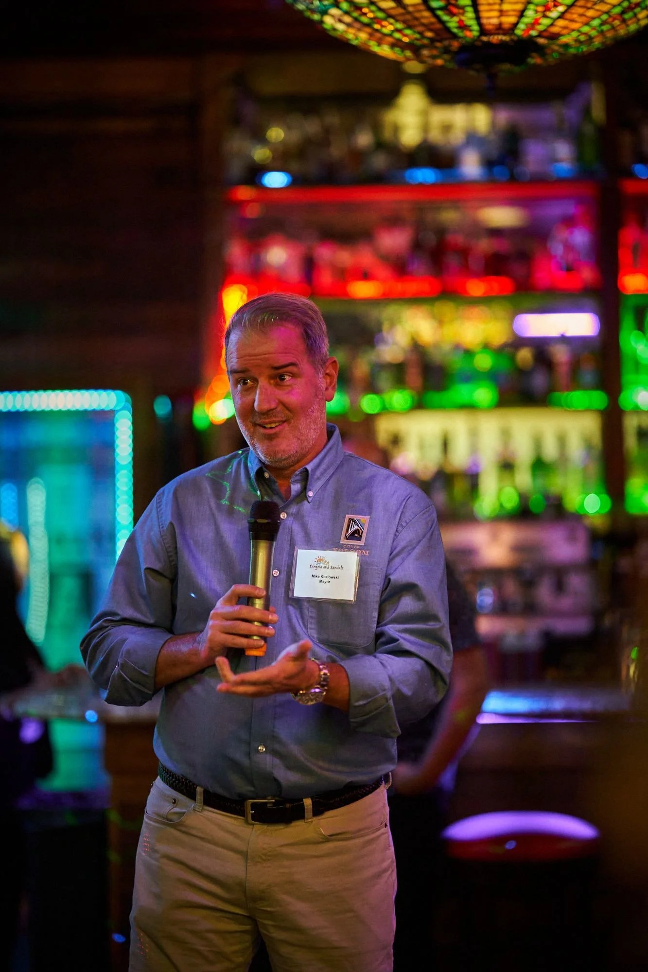 A man in a blue shirt holding a microphone, standing in a dimly lit entertainment or bar setting with colorful neon lights in the background.