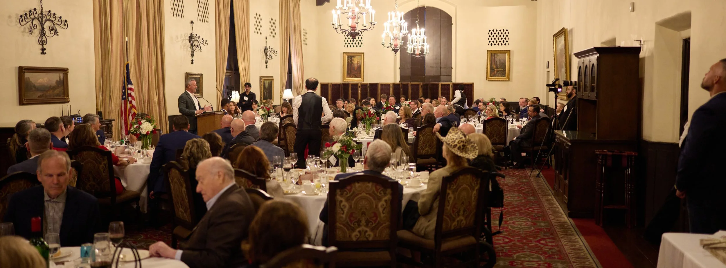 A formal banquet hall with round tables, floral centerpieces, and guests seated and listening to a speaker at a podium. The room has high ceilings, chandeliers, paintings, and large windows with drapes.