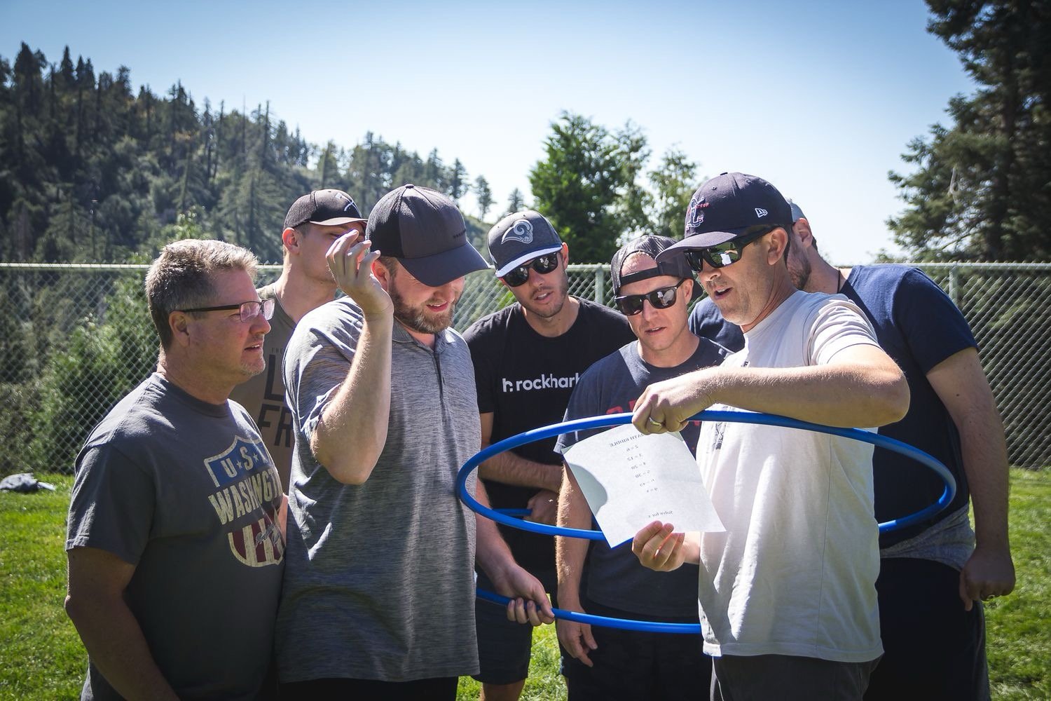 A group of seven men outdoors on a sunny day, gathered around a man holding a sheet of paper inside a blue hula hoop, with trees and a chain-link fence in the background.