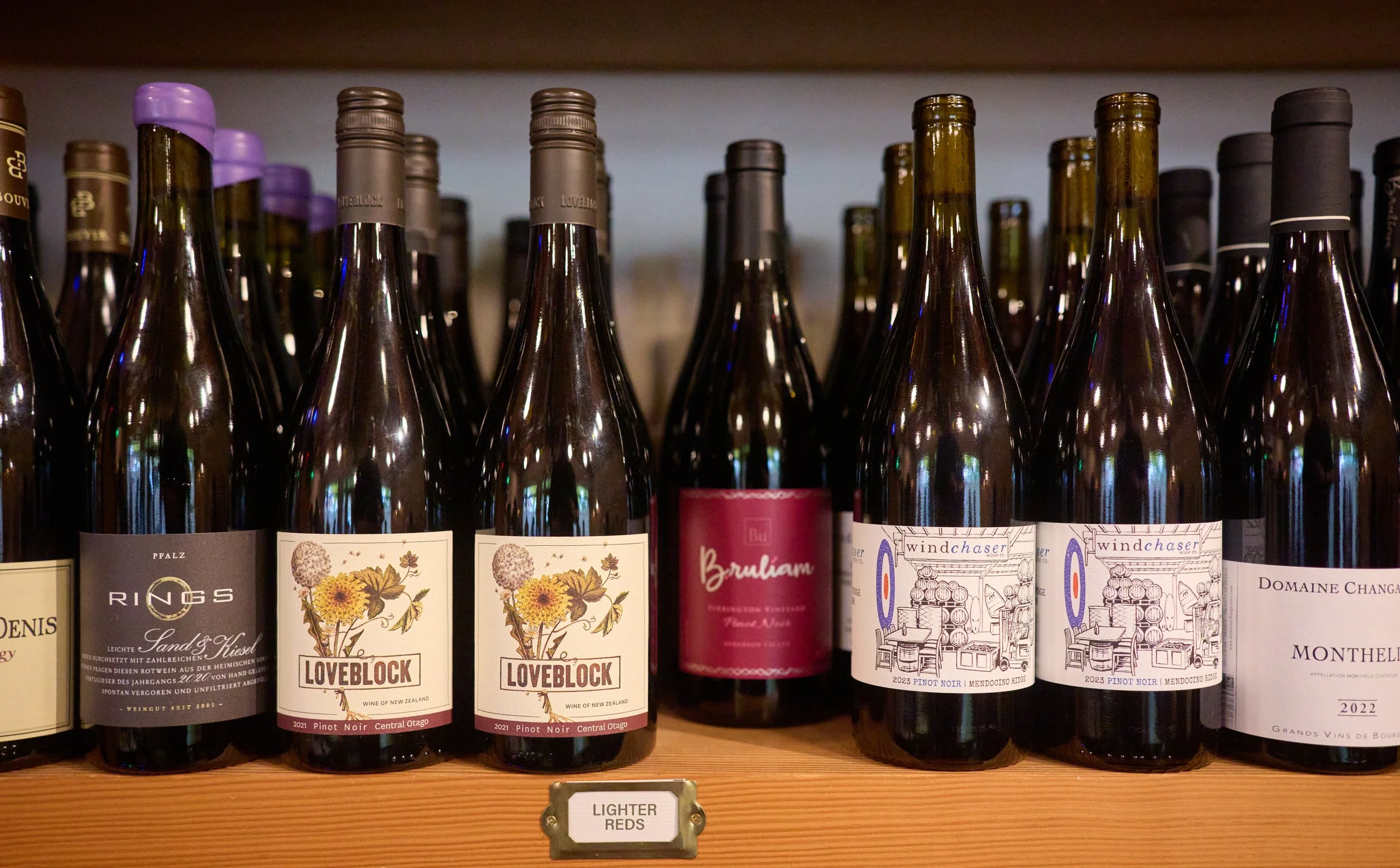 Several bottles of wine with colorful labels on a wooden shelf with a small sign reading "lighter reds" underneath.