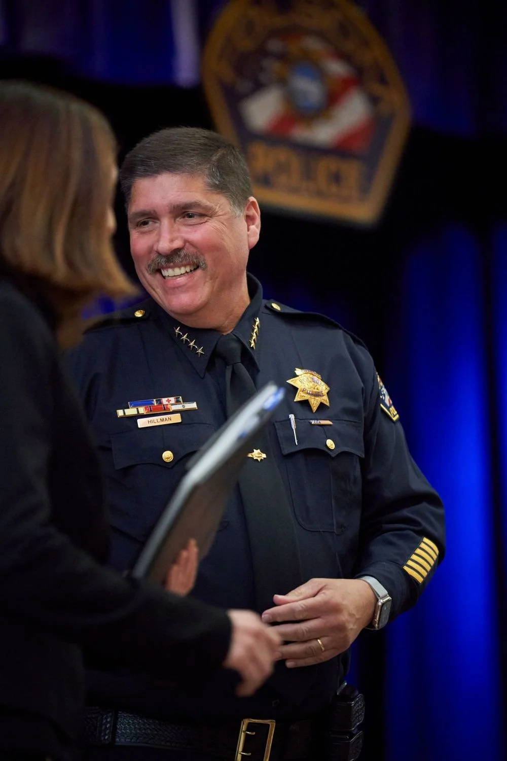A police officer smiling while talking to a woman holding a folder, with a police badge on his shirt and a badge behind them.