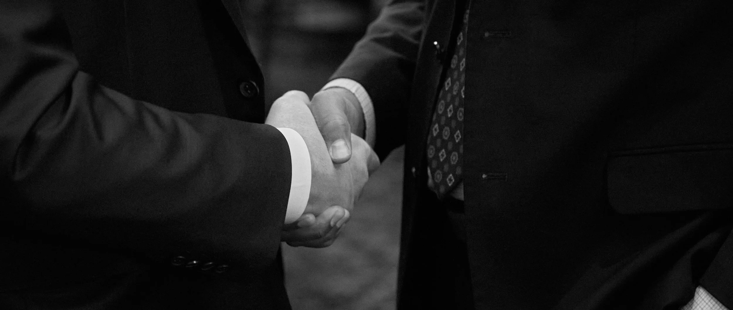 Close-up of two men in suits shaking hands, one wearing a patterned tie, in a professional setting.