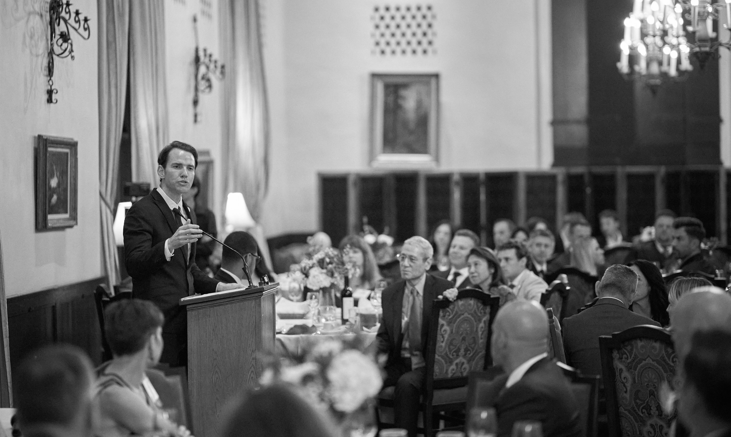 A man in a dark suit stands at a podium speaking to an audience in a large, elegant room. The audience is seated at tables with floral centerpieces, and there is a chandelier hanging from the ceiling. The room has decorated walls with paintings and c