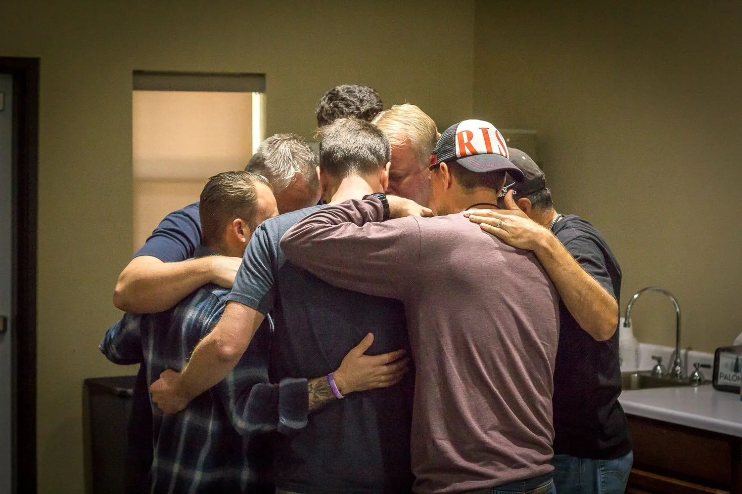 A group of people huddled together in a warm hug, heads bowed and arms around each other in an indoor setting.
