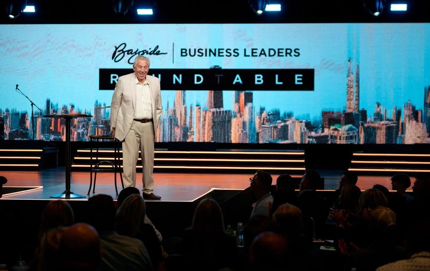 A man in a light suit speaking on a stage during a business leaders roundtable event, with a large digital screen behind him displaying the event's name.