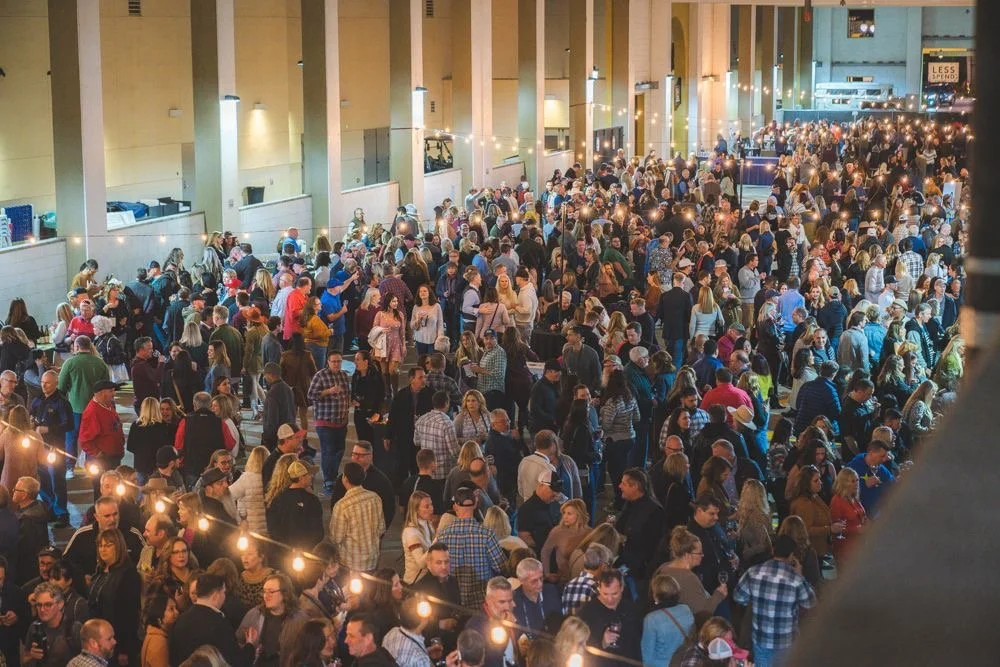 A large indoor gathering of people, possibly a concert or event, with string lights hanging overhead and a crowd of diverse individuals.