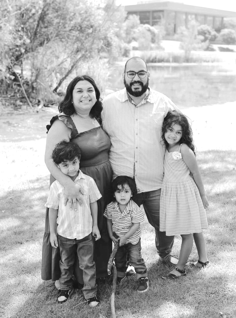 A family of five, including a woman, a man, and three children, standing outdoors on a grassy area near a body of water, smiling at the camera in black-and-white.