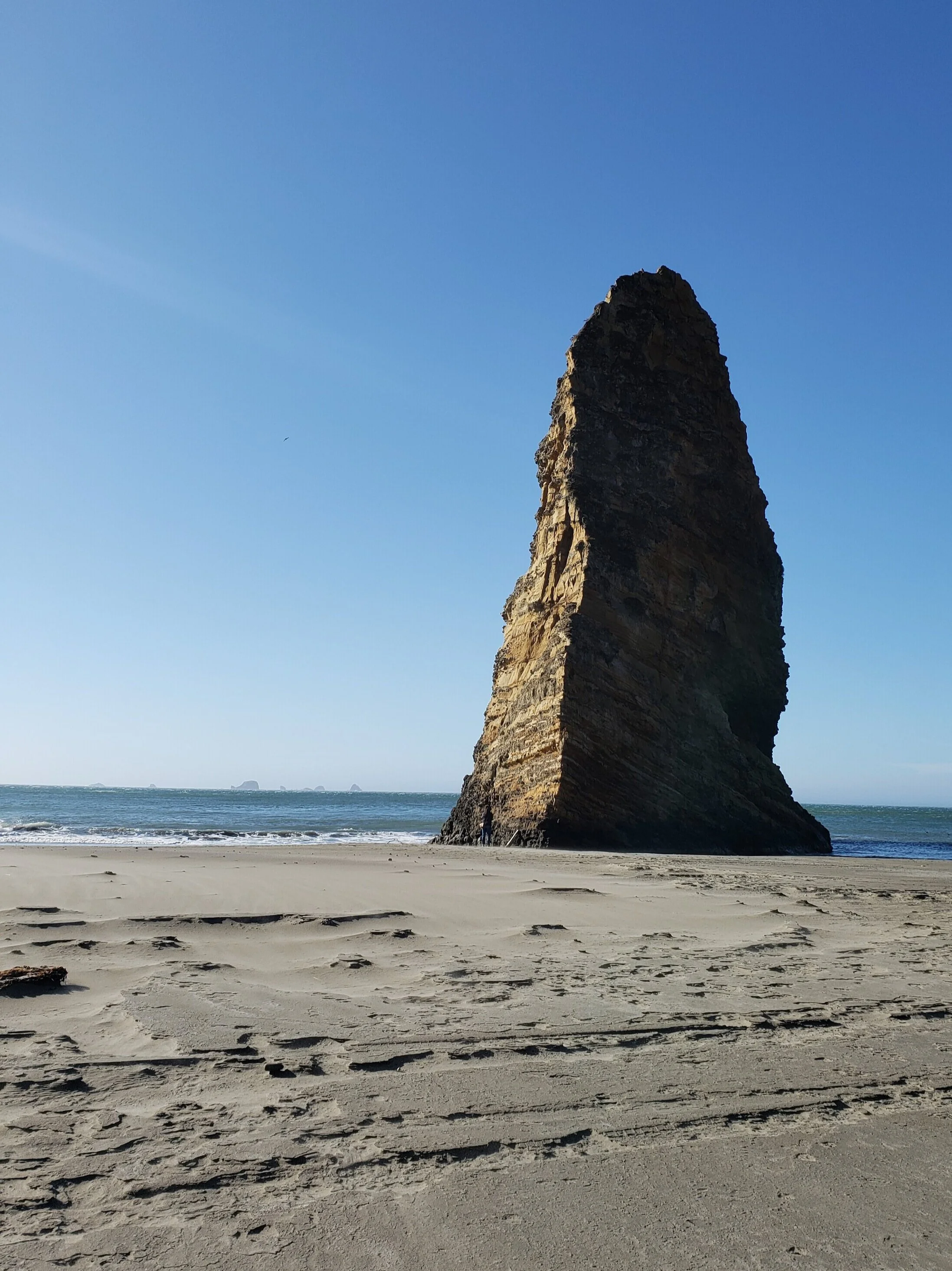 A tall, rocky sea stack rising from a sandy beach with waves in the background under a clear blue sky.