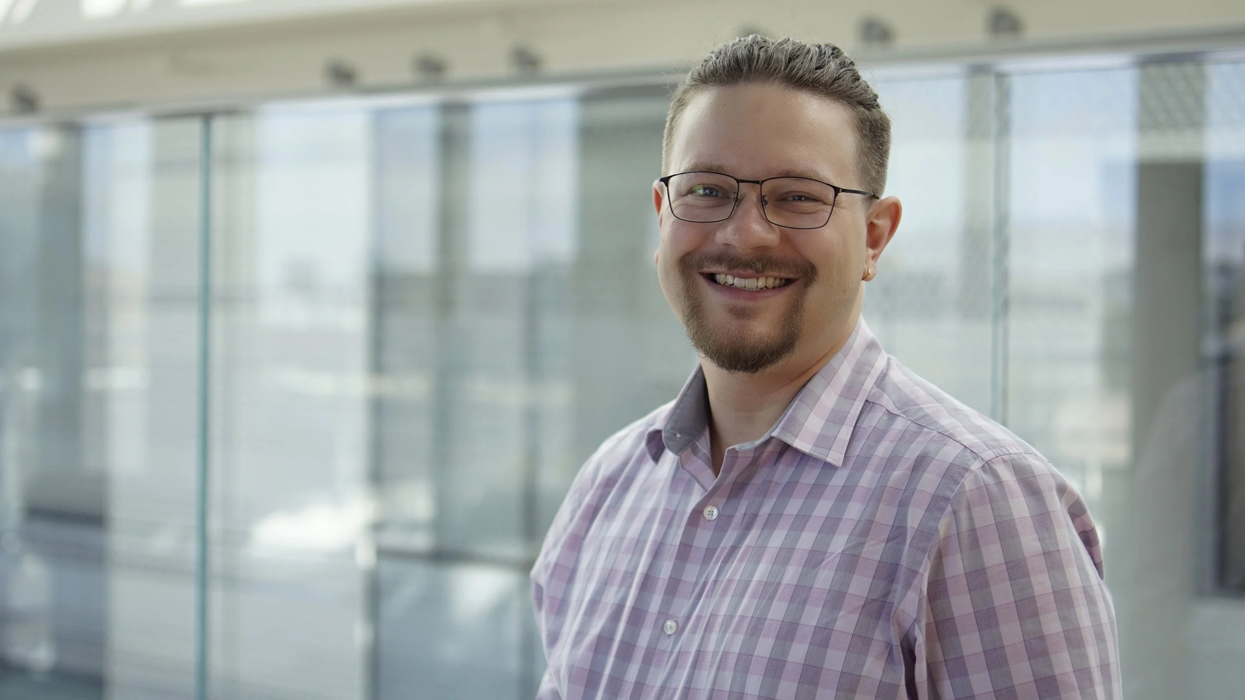 Man with glasses and a beard smiling in a professional setting with glass walls.