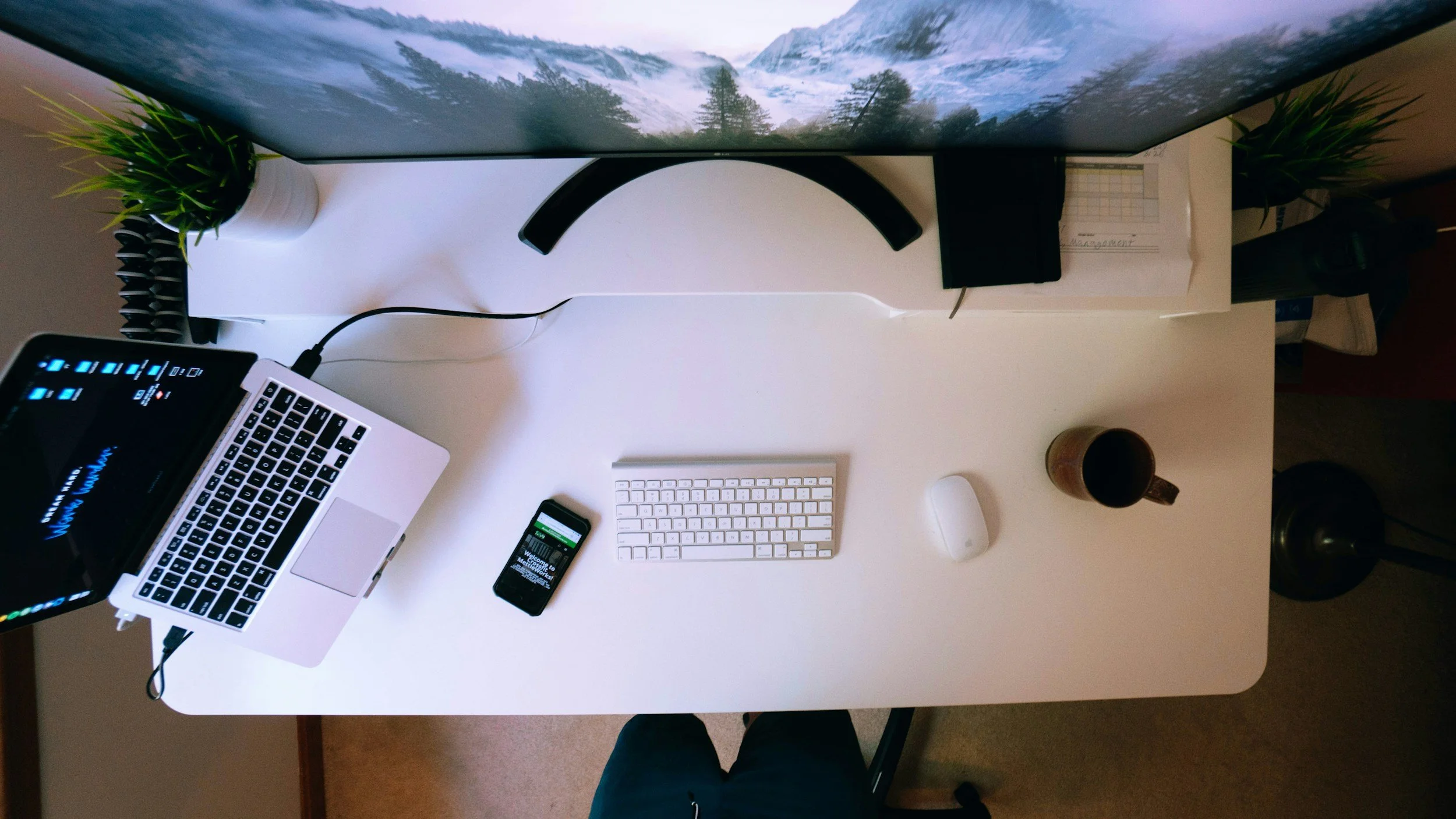 Top-down view of a clean, organized office desk with a laptop, smartphone, keyboard, mouse, two potted plants, a cup of coffee, and a large monitor showing a mountain landscape.