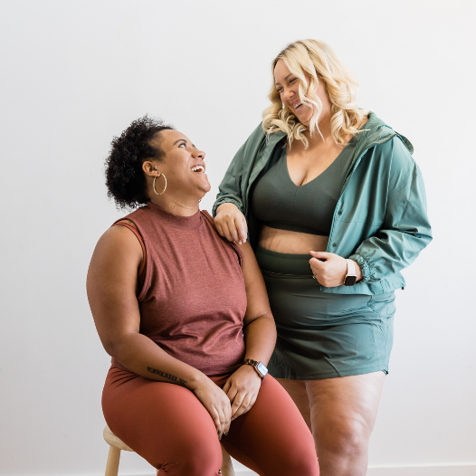 Two women smiling and looking at each other, one sitting on a stool wearing a sleeveless rust-colored top and matching pants, the other standing next to her in a green jacket and shorts, against a plain white wall.
