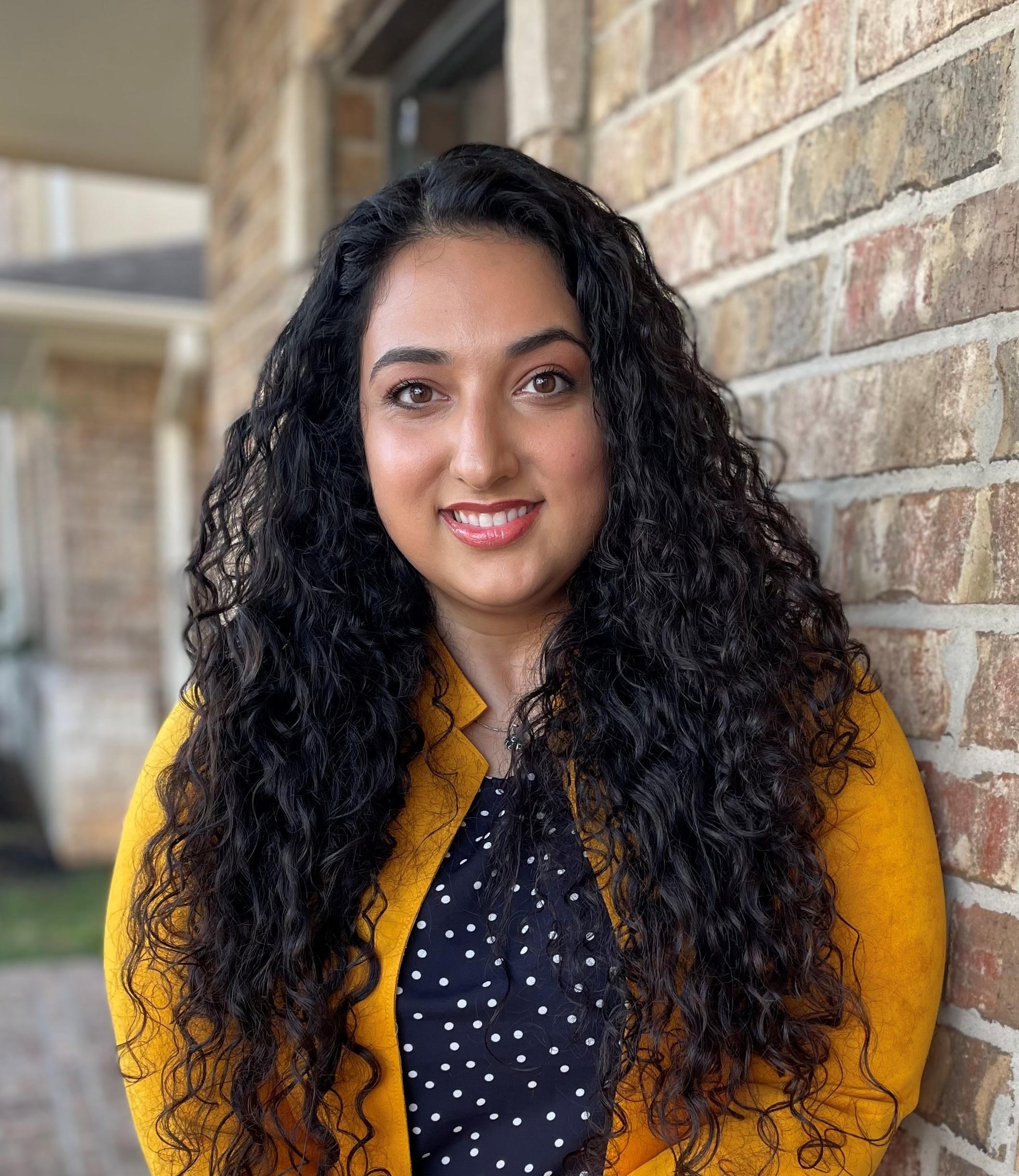 Yalda Ahmadi, LPC with long, curly black hair smiling in front of a brick wall, wearing a yellow blazer over a navy blue polka-dot shirt.