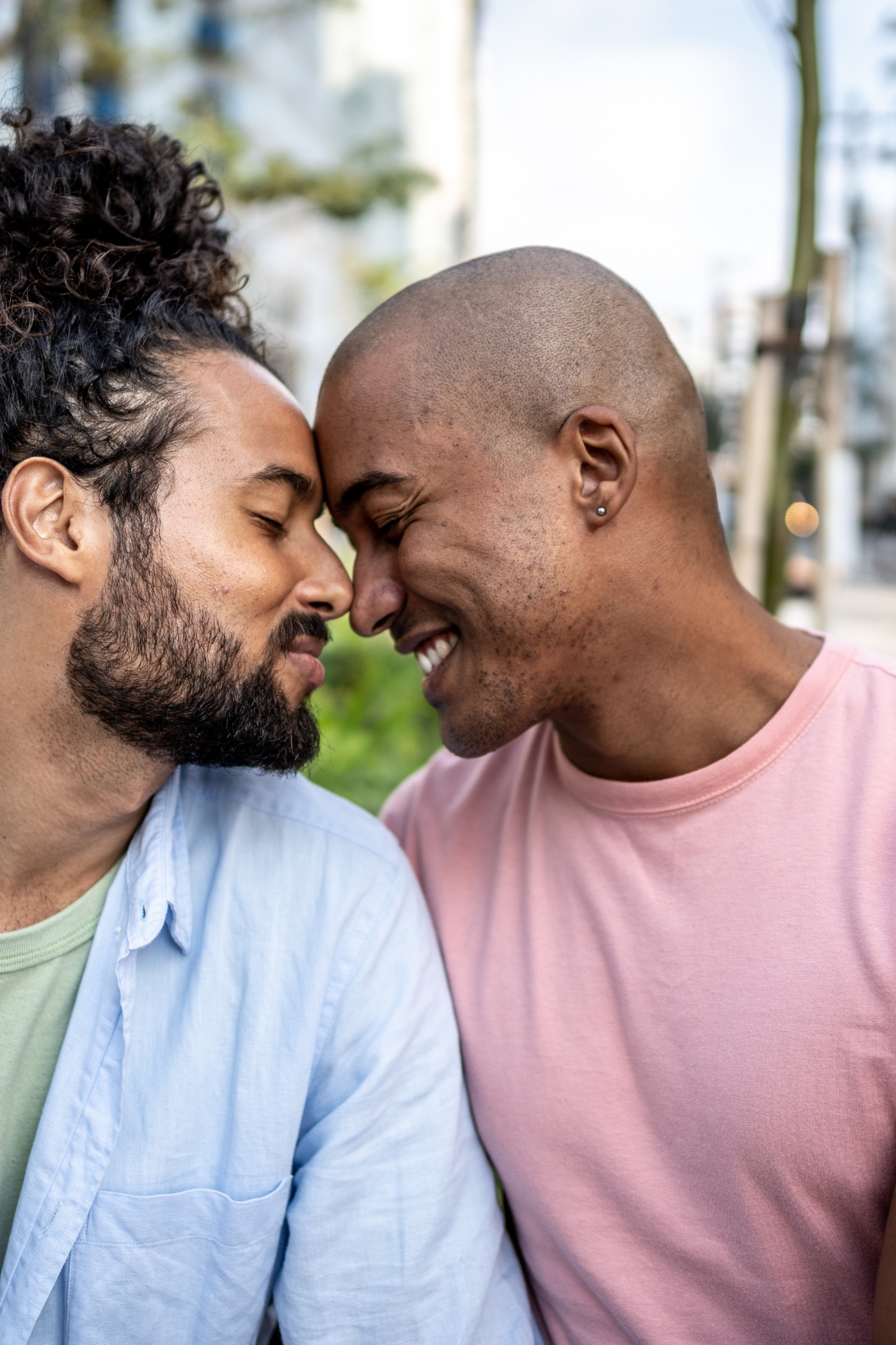 Two men with closed eyes touching foreheads and smiling, outdoors with blurred background of trees and buildings.