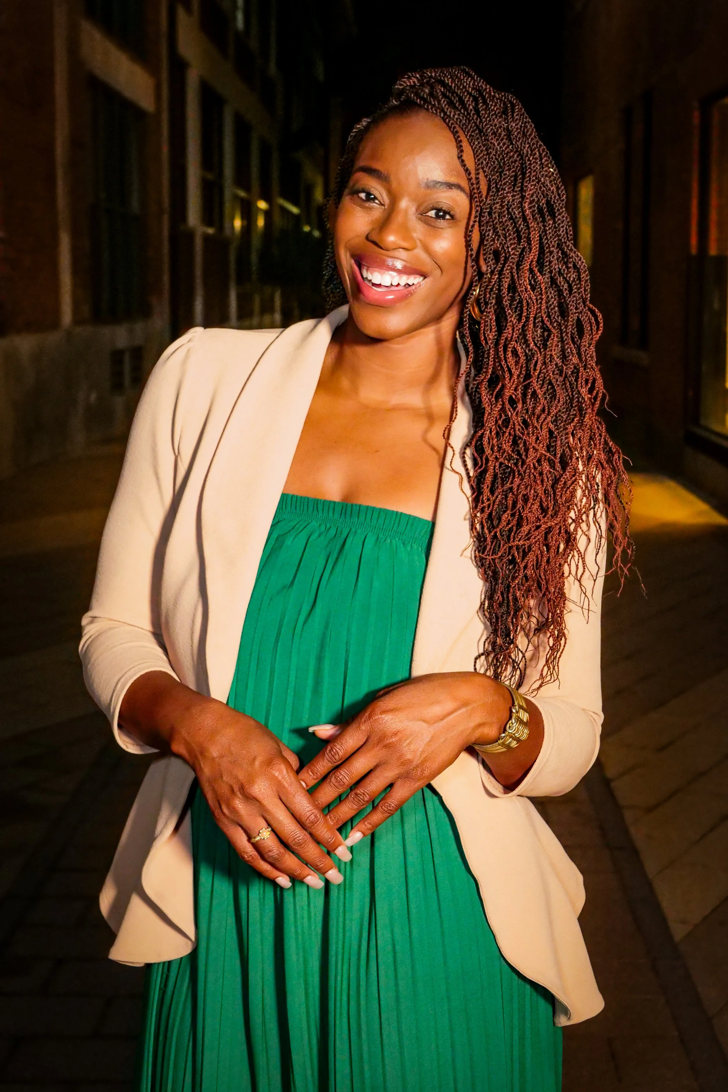 Phylica Bess, LPC-Associate with long, braided hair smiling at the camera, wearing a beige blazer over a green dress, with a gold watch and rings, standing in front of a dark city street background at night.