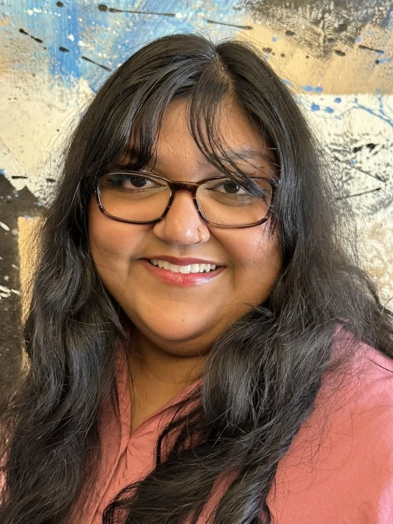 Nawaal Ahkwand smiling woman with long, dark hair, glasses, and a nose piercing wearing a pink top, standing in front of a textured wall with blue, black, and cream-colored paint.