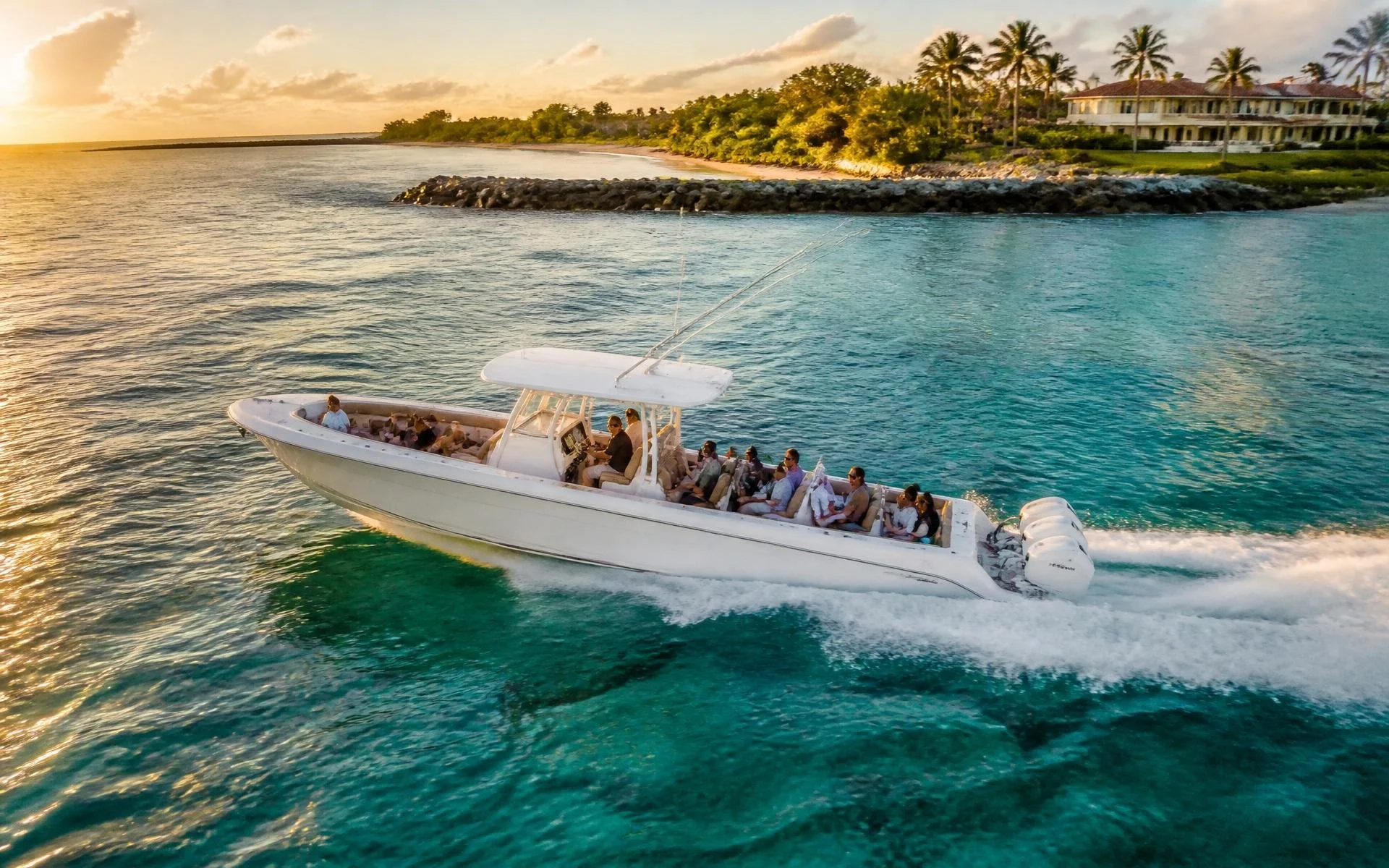 A group of people on a white boat with outboard motors, sailing in clear blue water near a tropical island with palm trees and large house, during sunset.