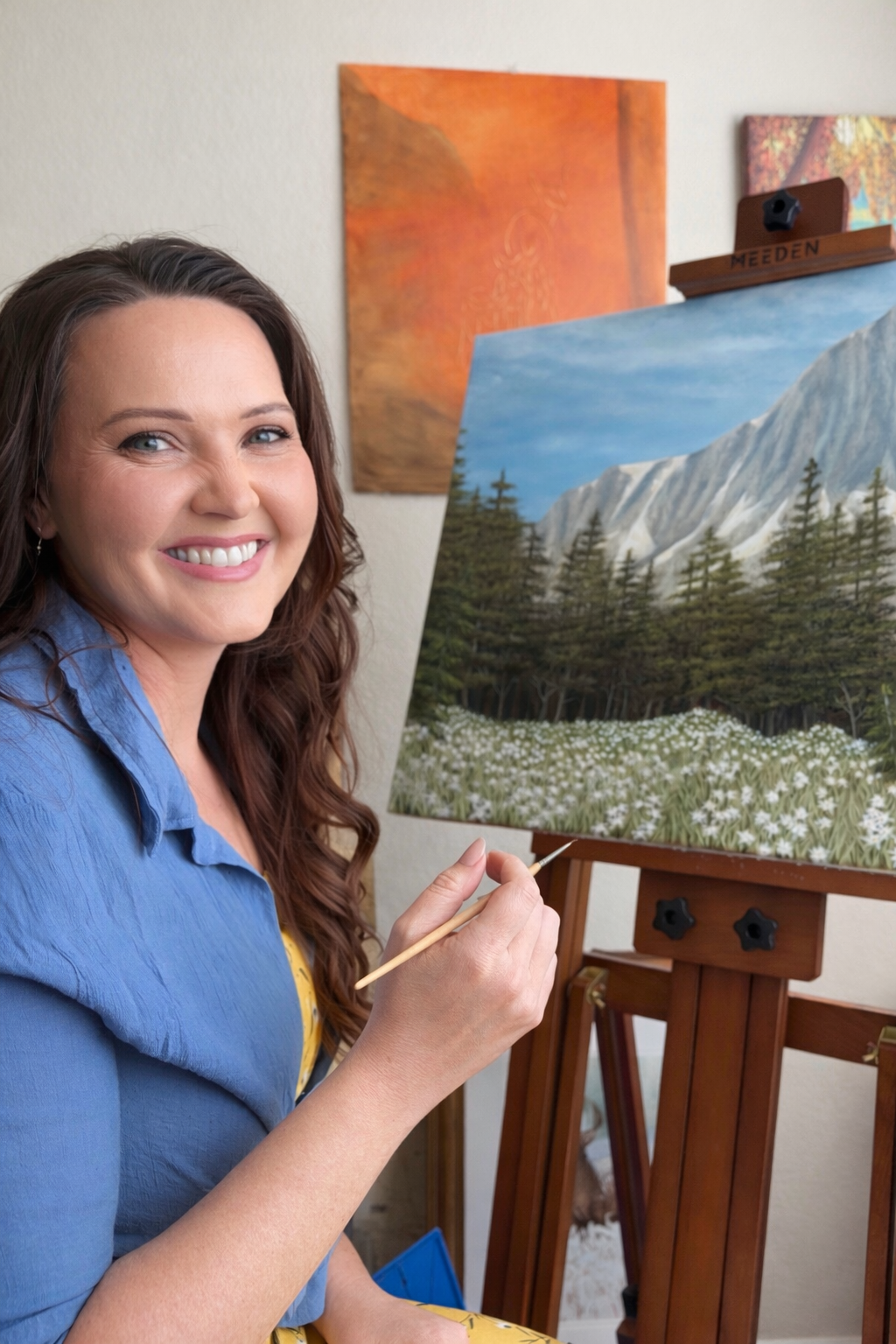 A woman smiling while painting a landscape of mountains, trees, and flowers on an easel.