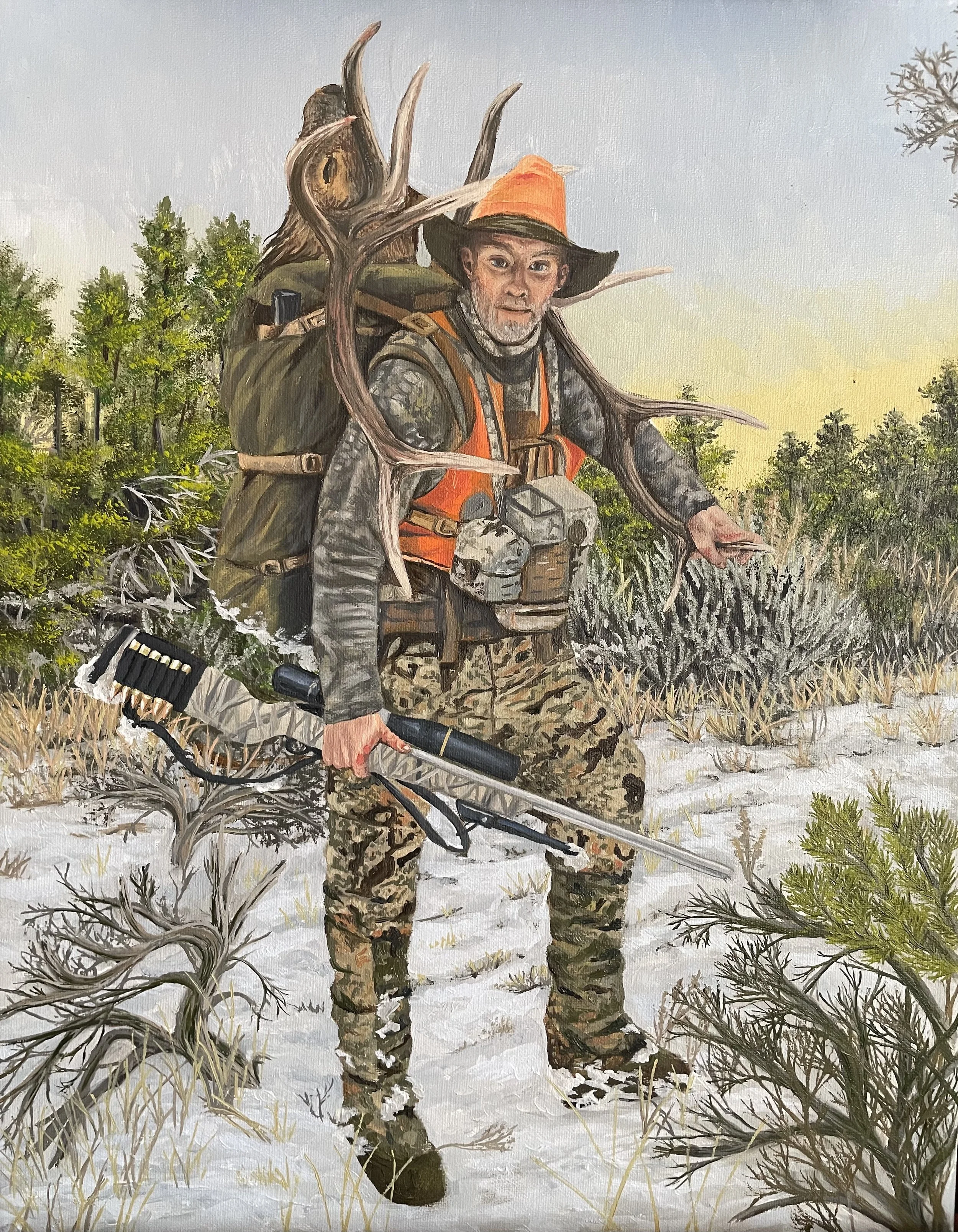 A man dressed in camouflage pants, a dark jacket, and an orange hat is walking through a snowy landscape while carrying a large backpack with orange and green colors. He has a beard and is holding a ski pole in his right hand. There are leafless shrubs and small trees around him, and the sky appears to be during sunset or sunrise.