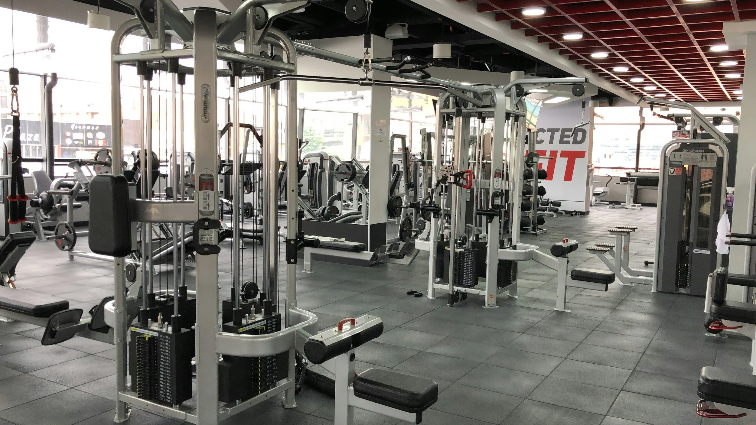 Inside a modern gym with various exercise machines, free weights, and equipment. Large windows let in natural light, and a sign with red and white text is visible in the background.