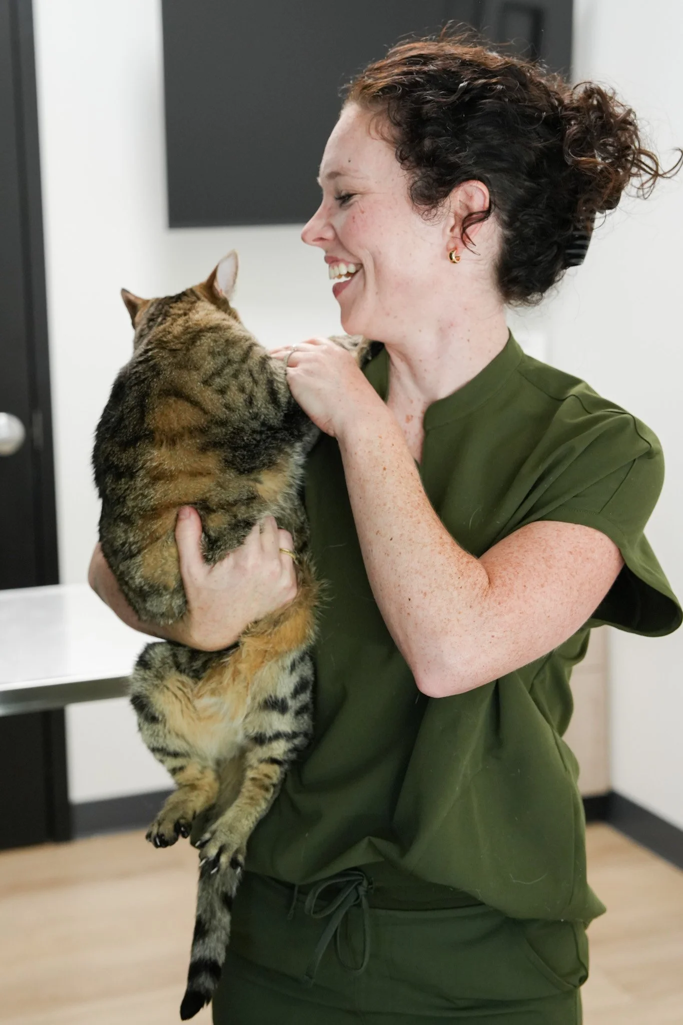 A woman with curly brown hair holding a tabby cat, smiling at the cat in an indoor setting.