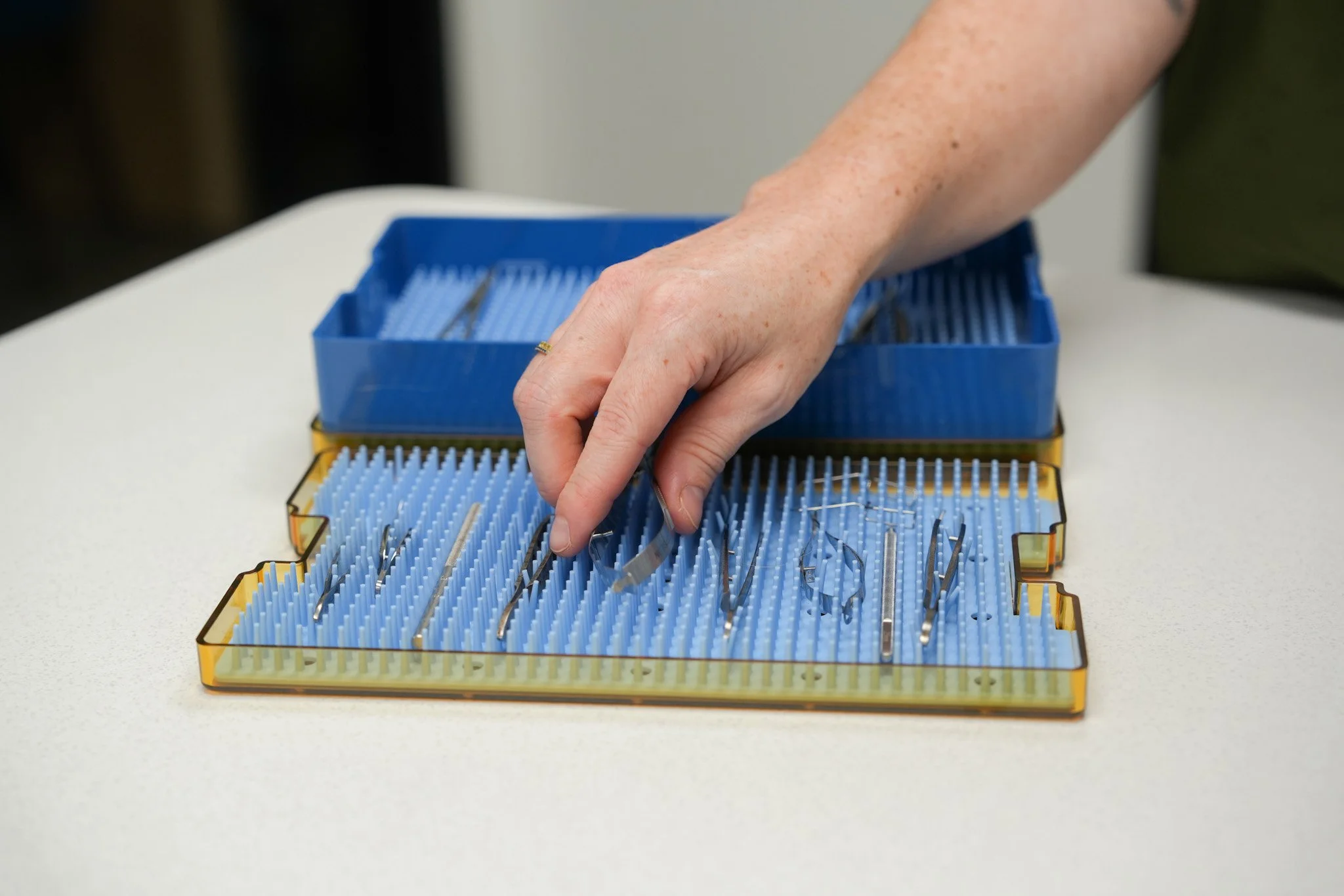 A person reaching into a blue surgical instrument tray filled with metal medical tools on a white surface.