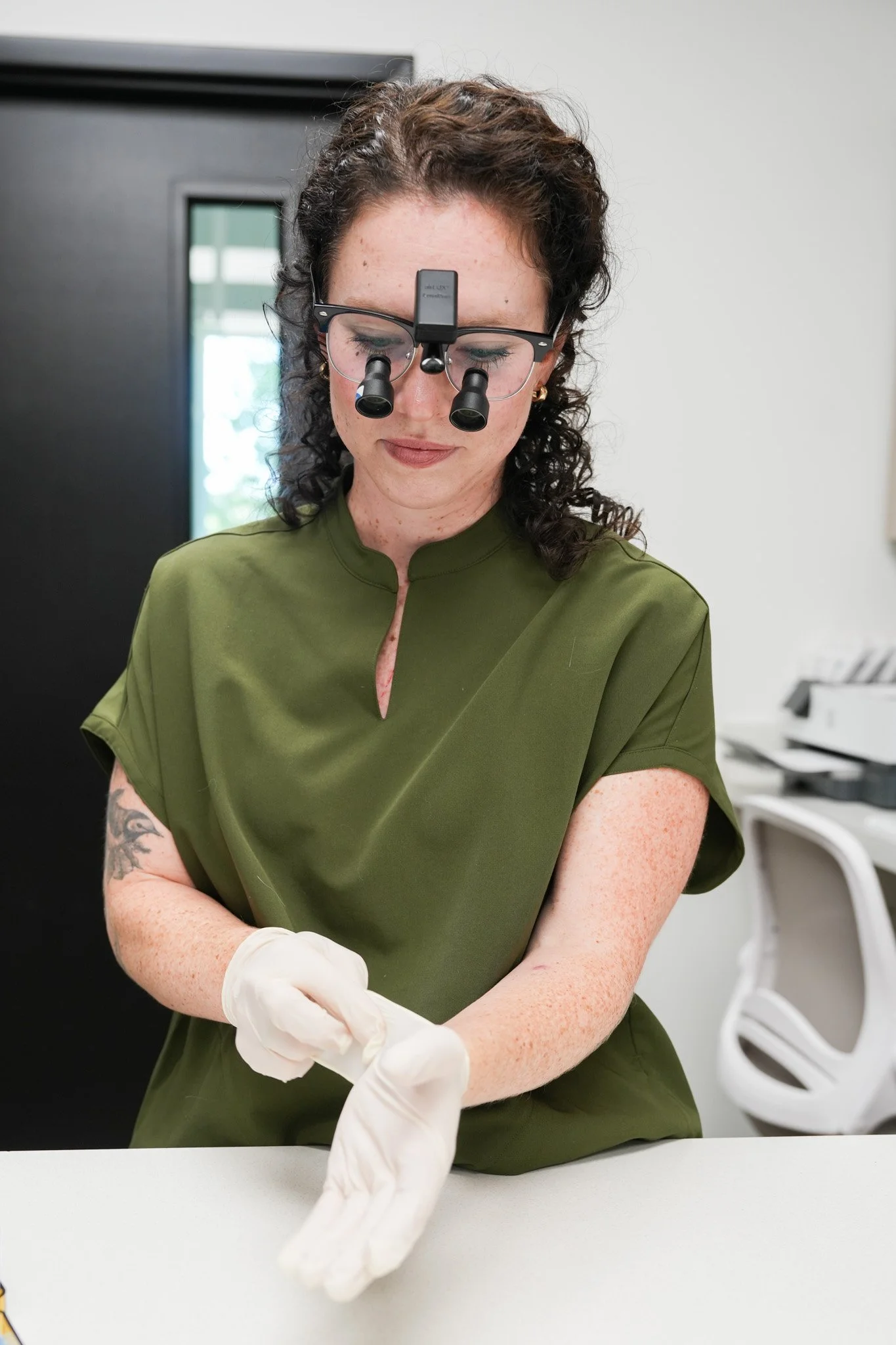 A woman in green scrubs wearing magnifying glasses with a headlight, examining or working on a task, with a doctor or medical professional's arm and gloved hand visible in the foreground.