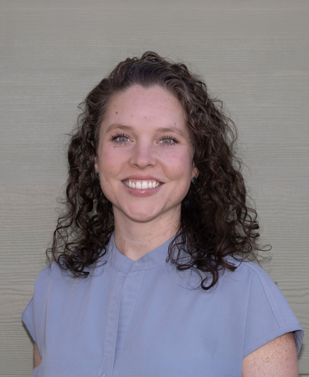 A woman with curly brown hair, blue eyes, and freckles, smiling and wearing a light blue collared shirt, standing in front of a textured beige background.