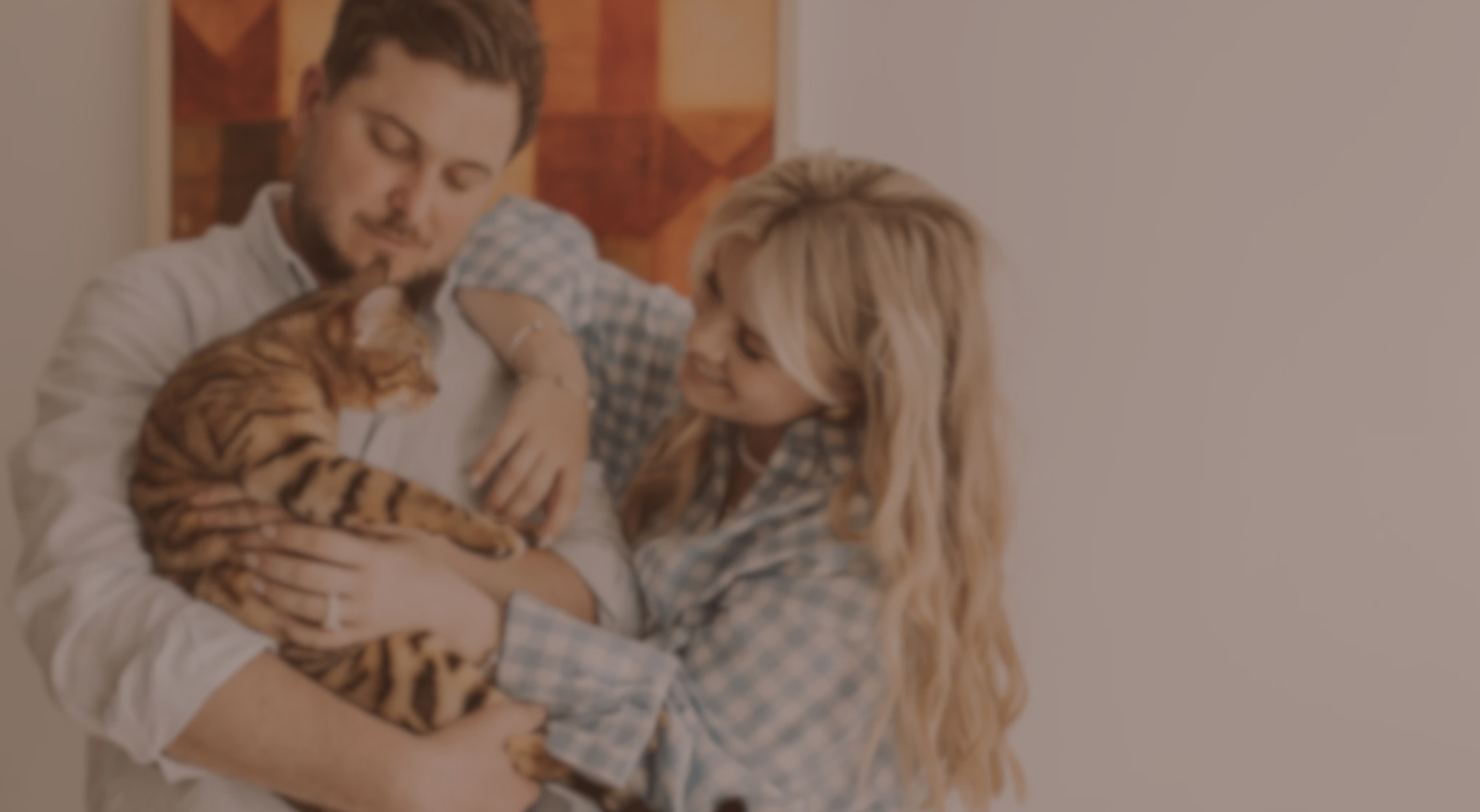 A man holding a Bengal cat with orange and black stripes, a woman leaning in affectionately, in a warmly lit room with a red and orange abstract painting on the wall behind them.