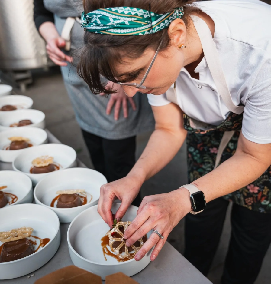 A woman with glasses and a green bandana prepares dessert bowls at a catering event, garnishing with a decorative fried lotus root slice.