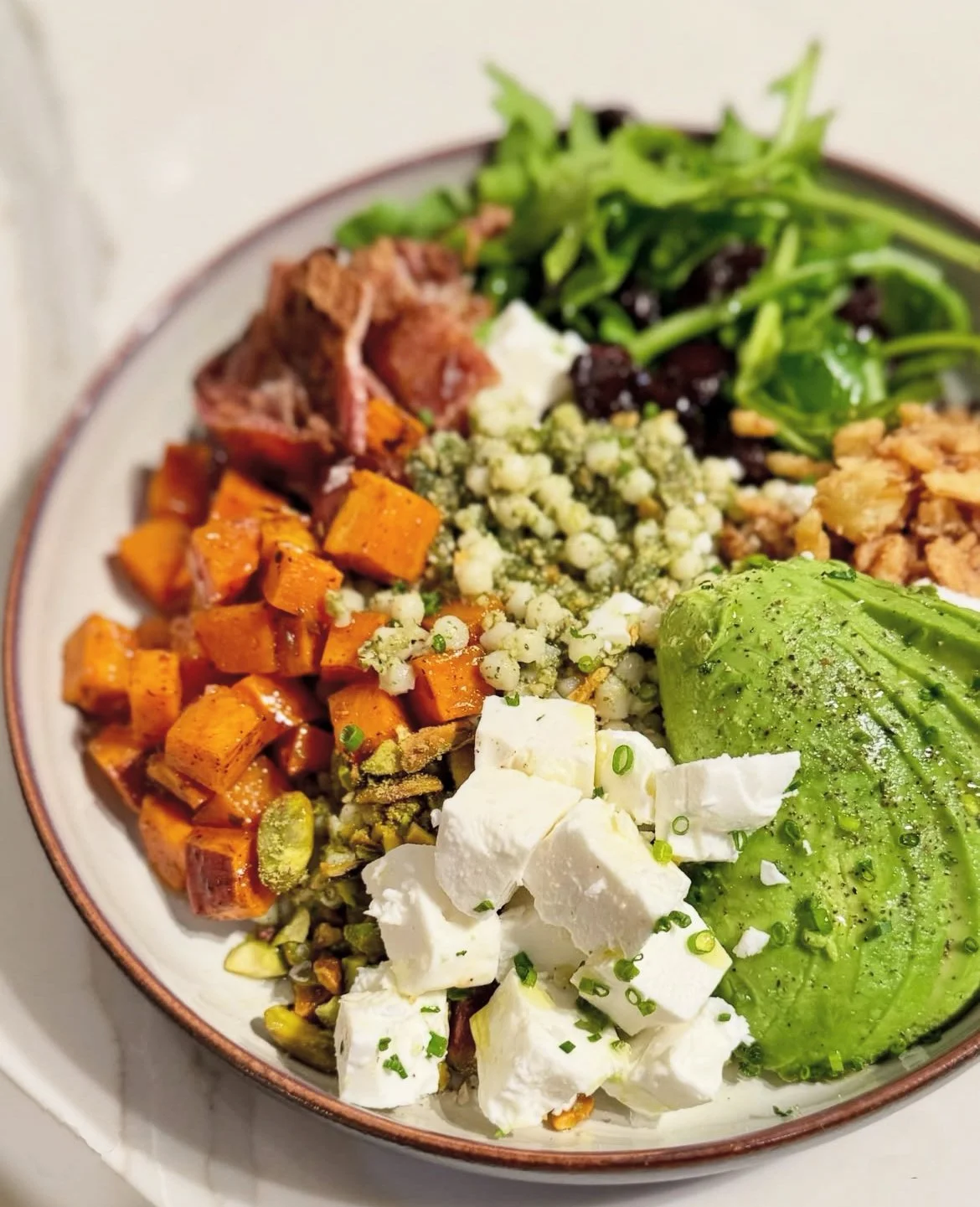 A bowl with roasted sweet potatoes, greens, crumbled cheese, sliced avocado, and assorted vegetables.